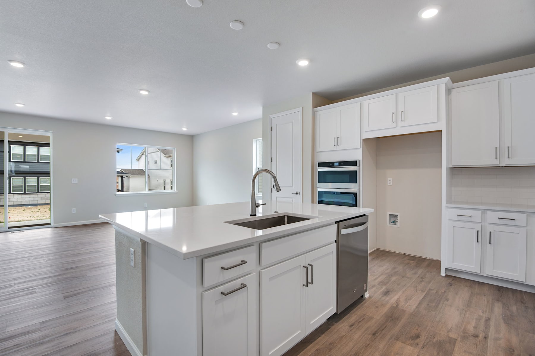 A modern, well-lit kitchen with white cabinets, a large island with a sink, and hardwood floors, set against a backdrop of windows and a bright, open living space.