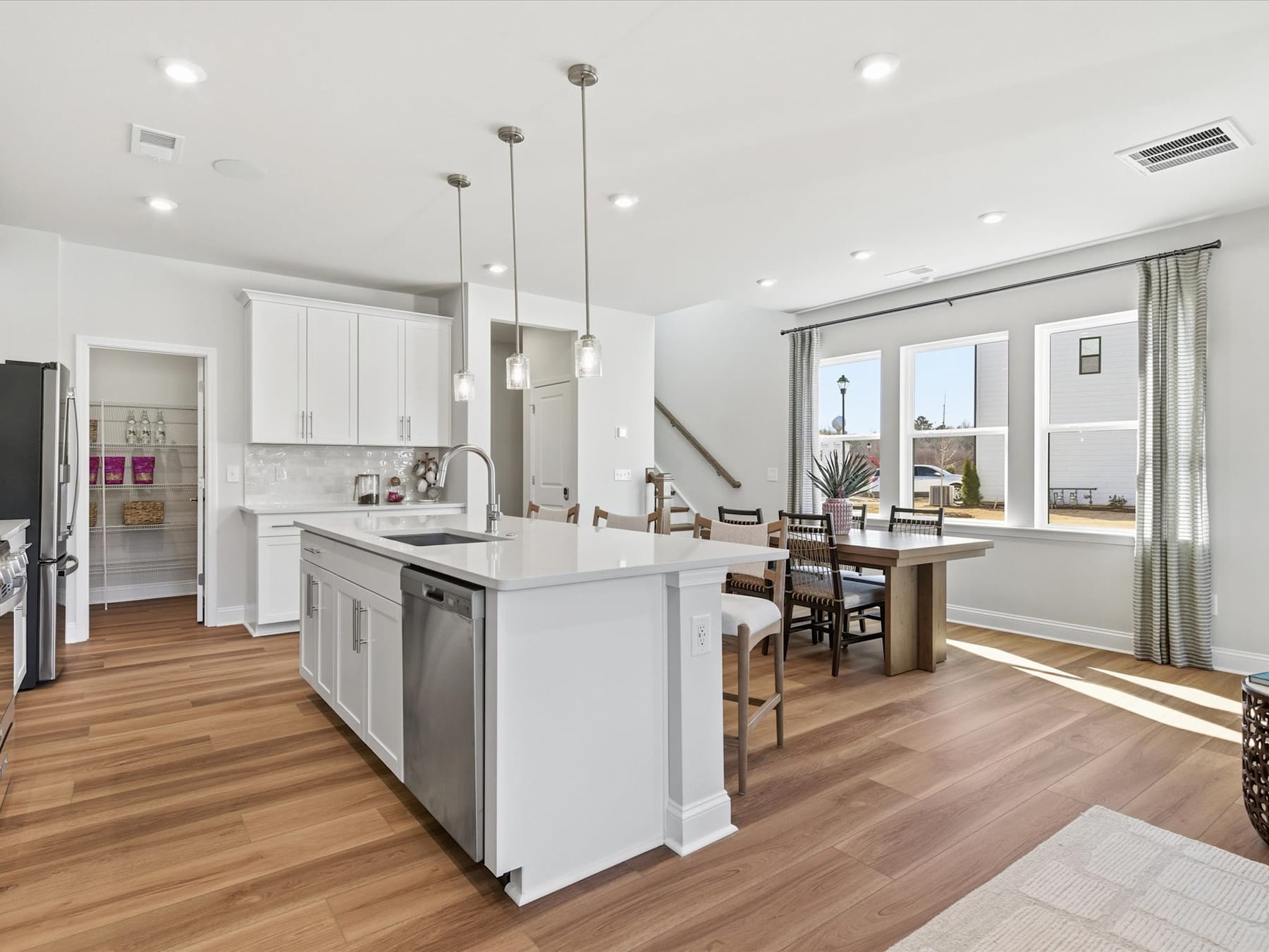 A modern, open-concept kitchen with white cabinets, stainless steel appliances, and a wooden dining table in the background, surrounded by large windows that let in natural light.