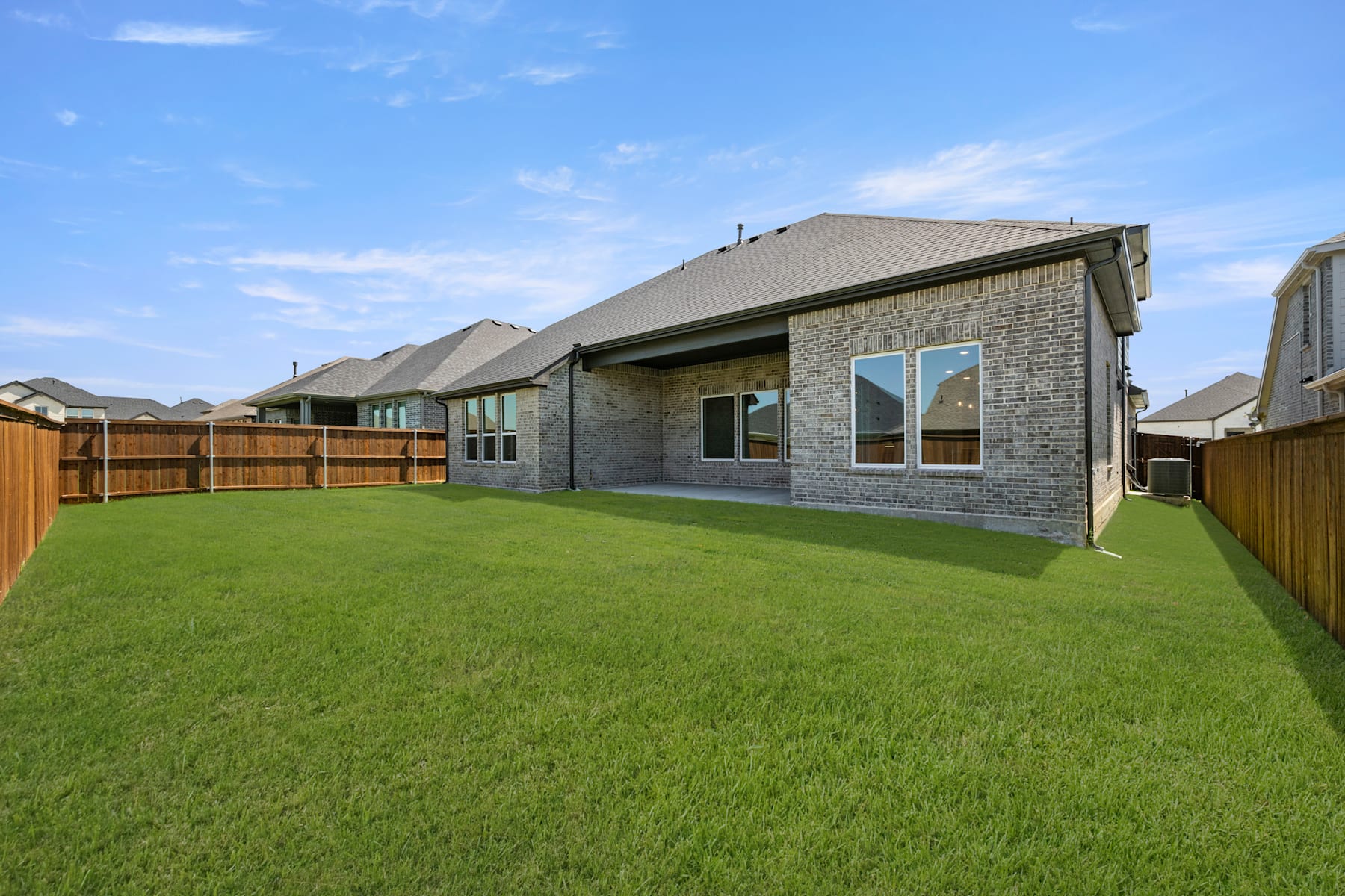 A well-manicured green lawn in the foreground leads to a modern brick house with a slanted roof and large windows in the background, set against a clear blue sky.