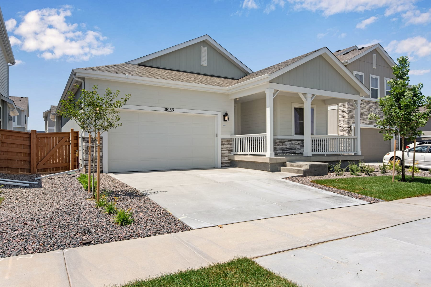 A well-maintained, two-story residential house with a paved driveway, a small lawn, and landscaping elements in the foreground, set against a clear blue sky with scattered clouds.