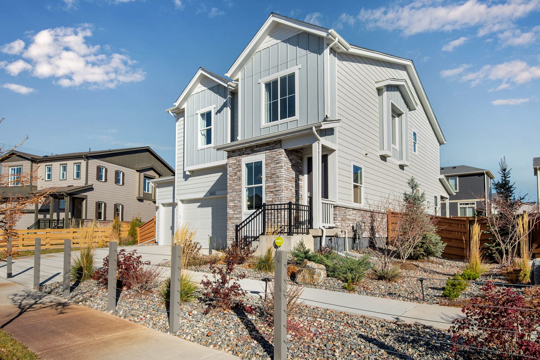 A modern, two-story house with a stone facade, surrounded by a landscaped yard with gravel and plants, set against a clear blue sky with scattered clouds.