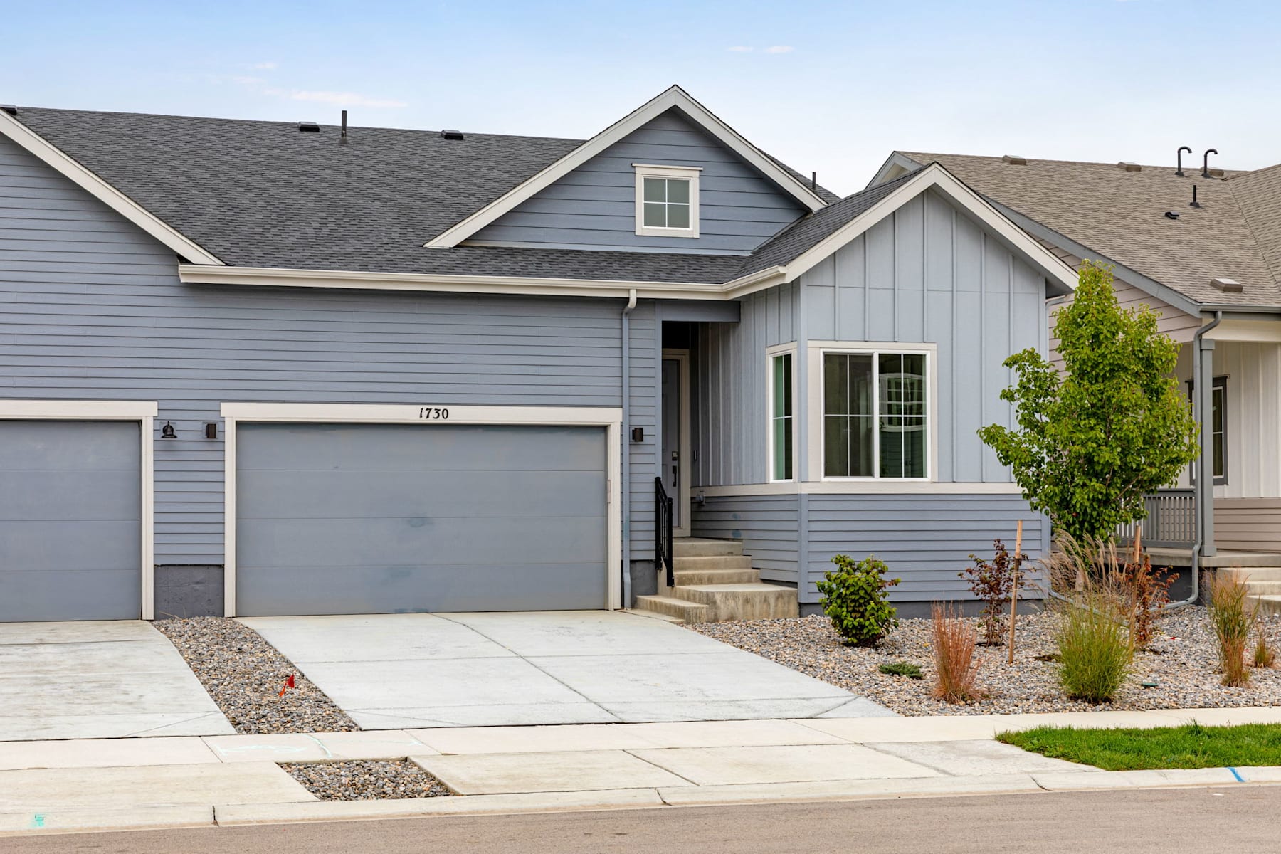 A modern, two-story gray house with a garage, surrounded by a well-maintained lawn and landscaping, stands against a clear blue sky.
