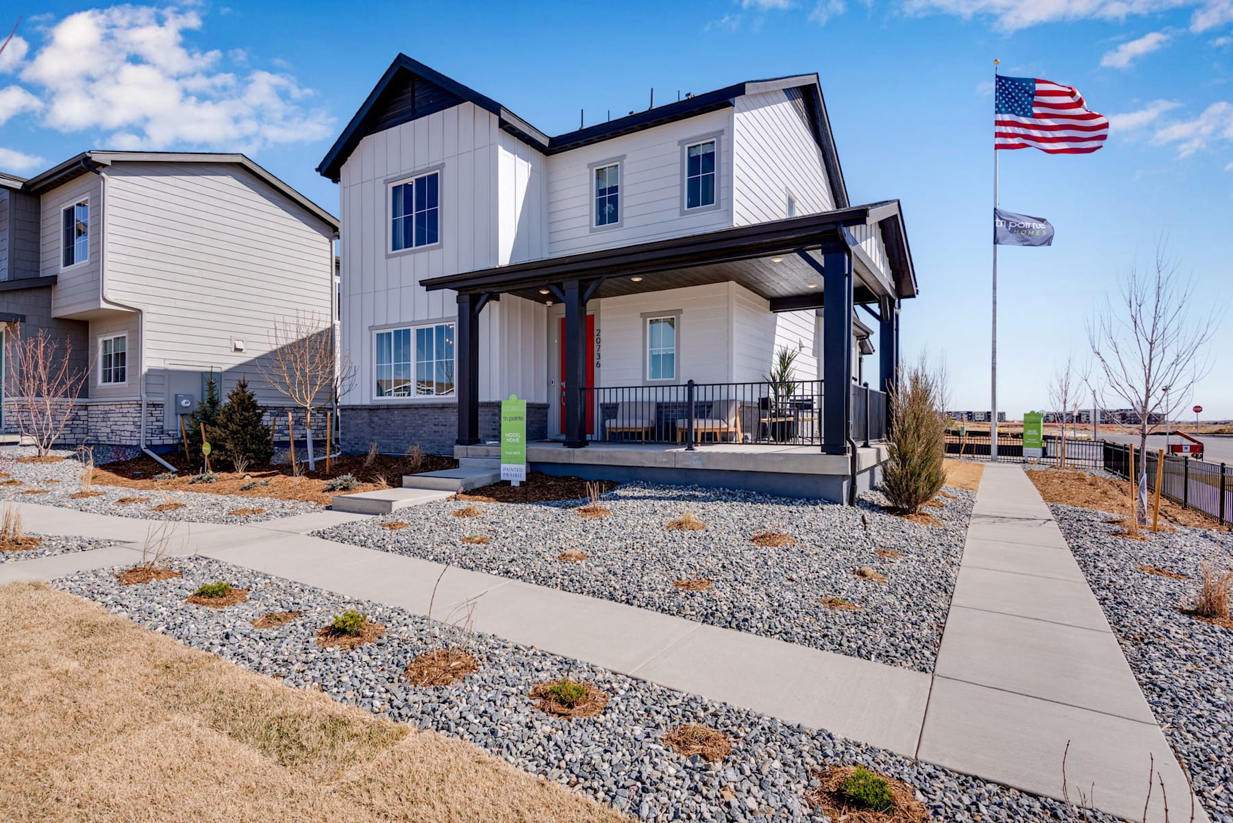 A modern, two-story white house with a covered porch, surrounded by a gravel pathway and landscaping, stands against a blue sky with scattered clouds, with an American flag flying in the foreground.
