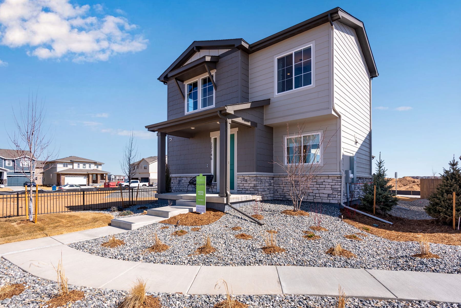 A modern two-story house with a gray exterior, surrounded by a gravel landscaping and a paved walkway, set against a clear blue sky with scattered clouds.