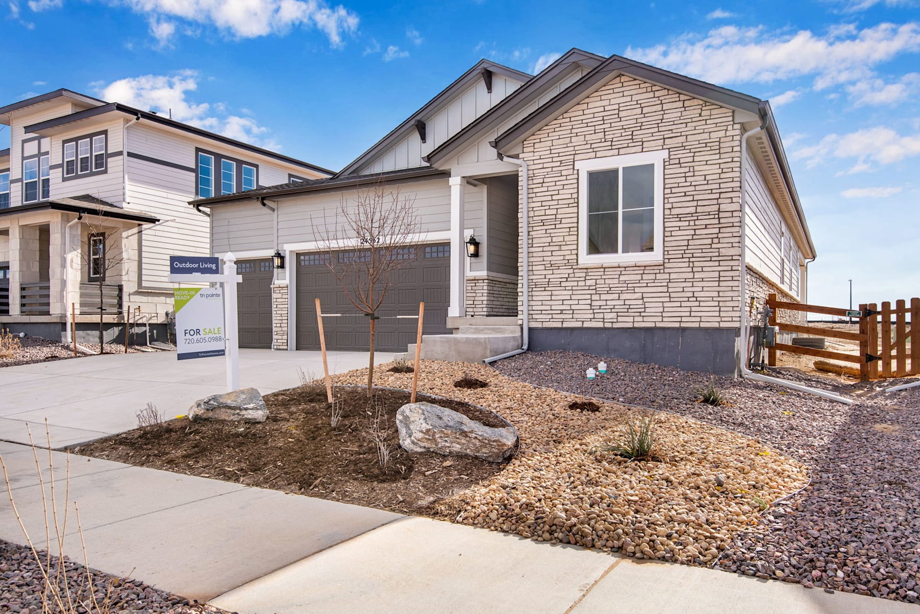 A modern two-story residential building with a stone exterior, surrounded by a landscaped yard with gravel and rocks in the foreground, set against a blue sky with scattered clouds.