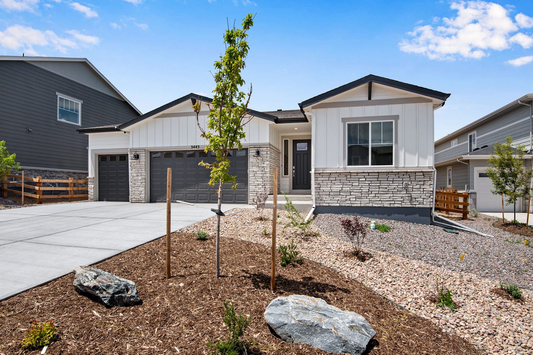 A modern, two-story house with a stone exterior, surrounded by a landscaped yard featuring a young tree, rocks, and mulch in the foreground.