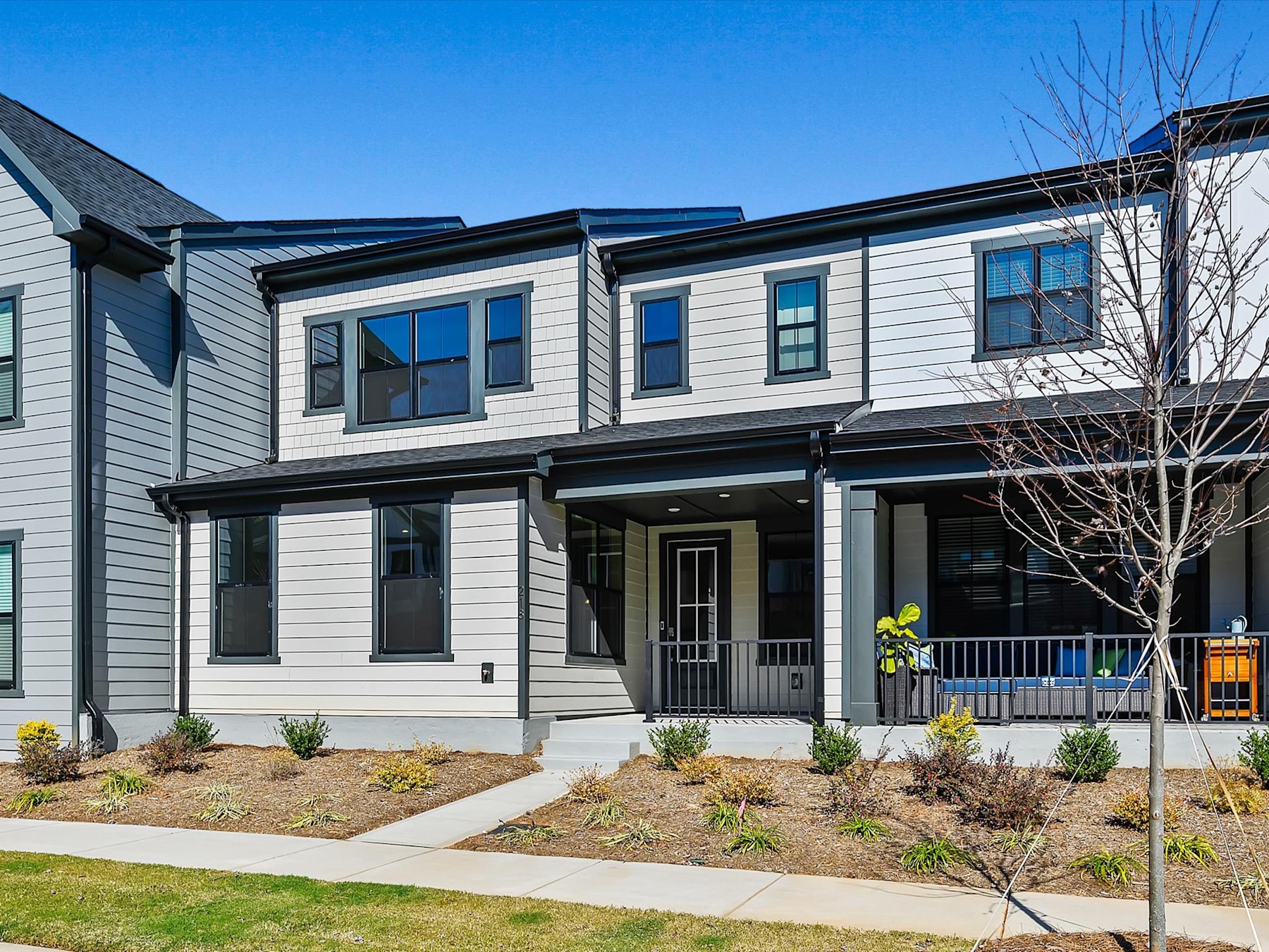 A modern two-story townhouse with a porch, surrounded by landscaped yards and trees against a clear blue sky.