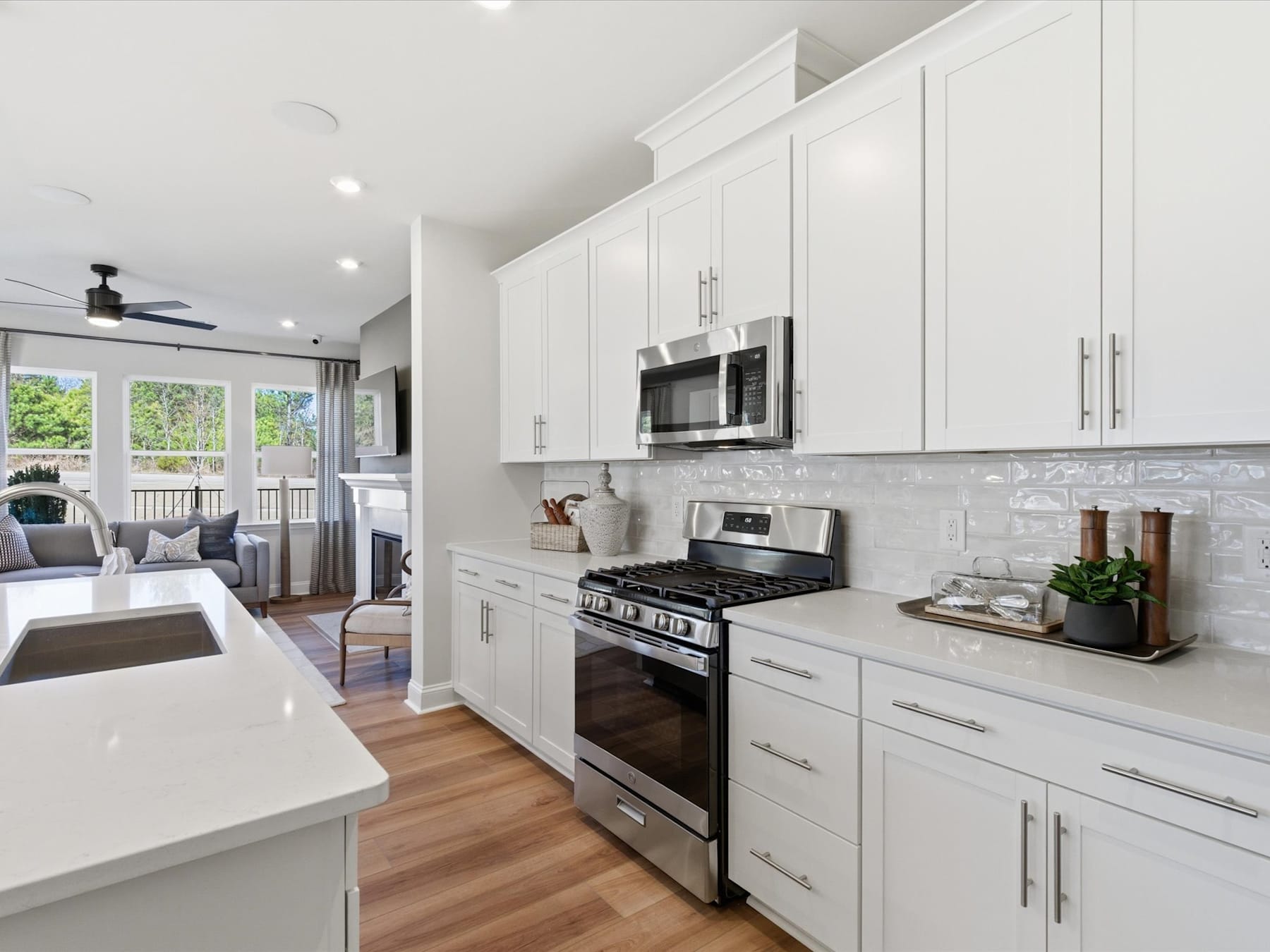 A modern, bright kitchen with white cabinets, stainless steel appliances, and a wooden floor, with a view of a living room area in the background.