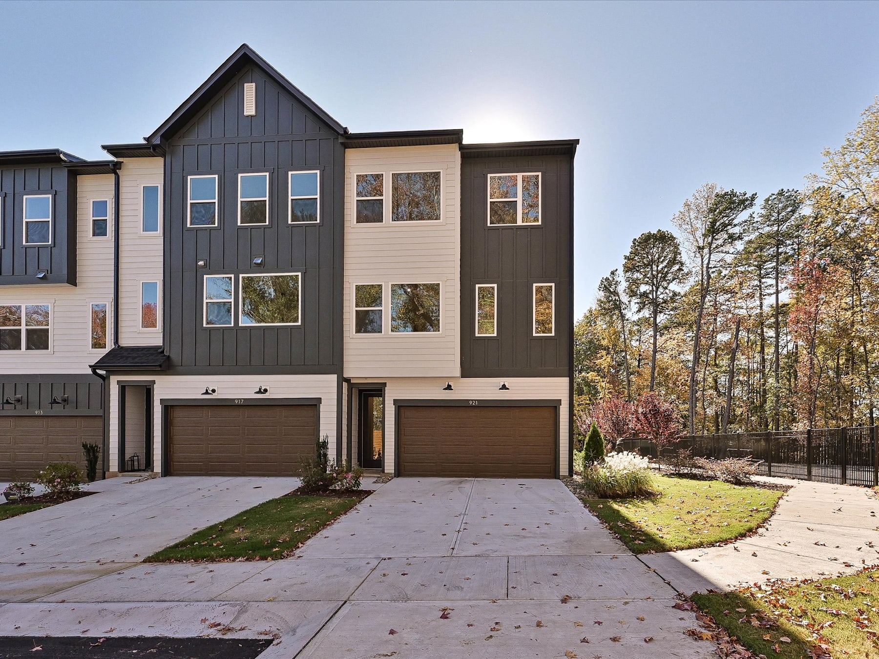 A modern, multi-story townhouse with a garage and a well-maintained lawn, surrounded by trees and a clear blue sky in the background.