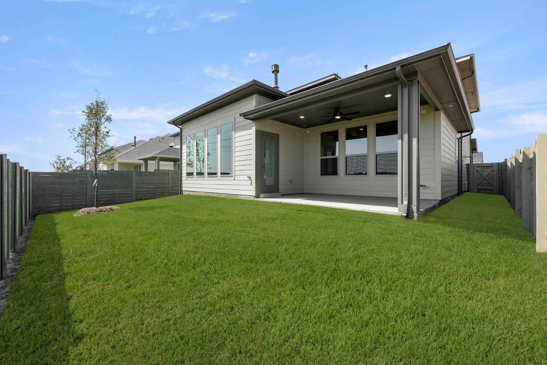 A modern, single-story house with a large grassy yard and a wooden fence in the foreground, set against a clear blue sky.
