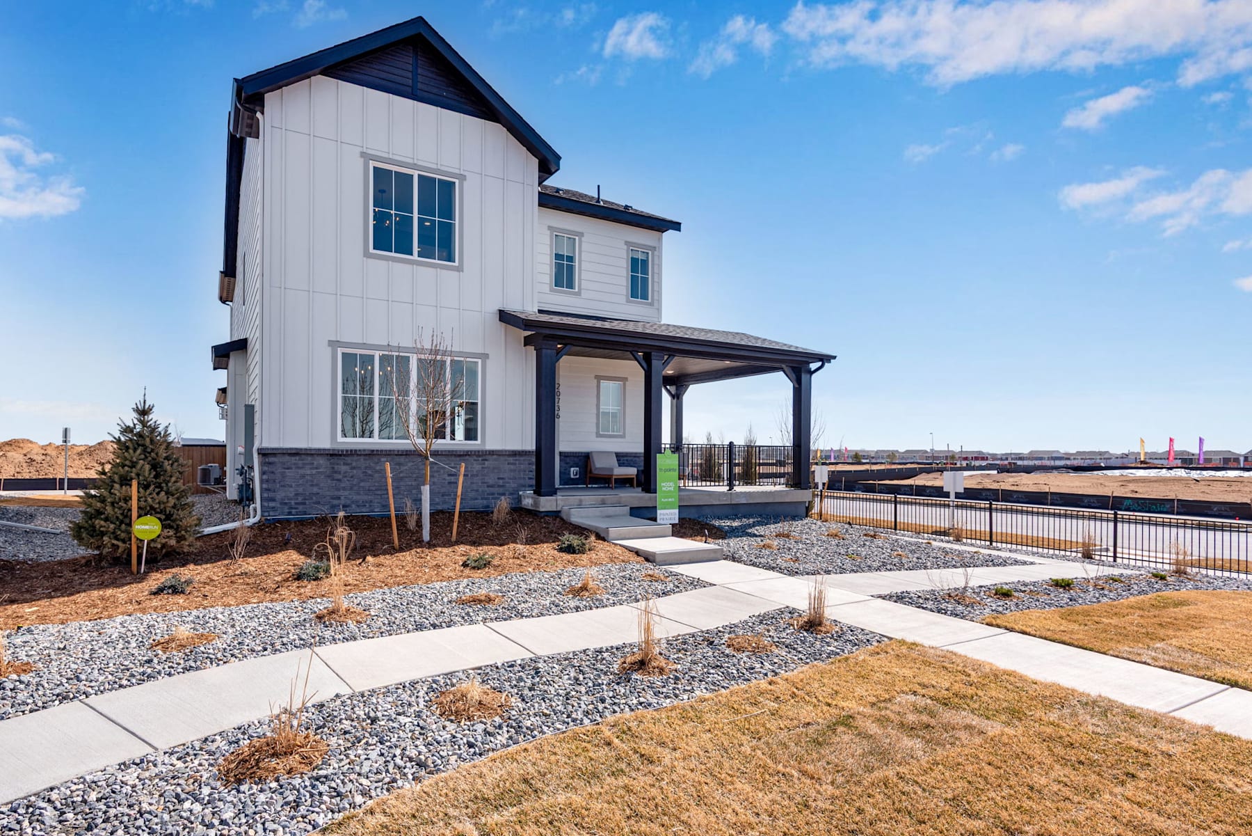 A modern, two-story white and gray house with a covered porch sits on a landscaped lot with a gravel path leading to the front door, surrounded by a clear blue sky with scattered clouds.