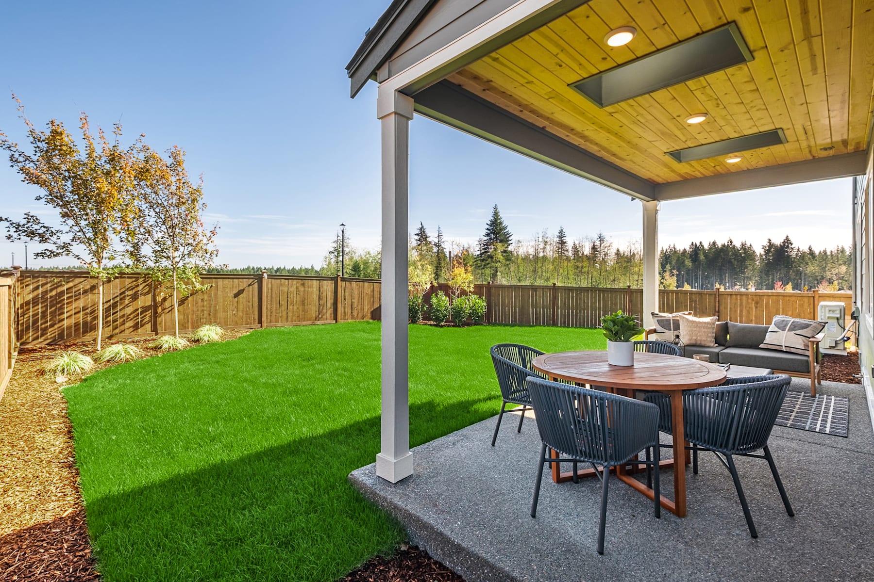 A well-manicured backyard with a covered patio, a lush green lawn, and a wooden fence surrounding the property, with a forested area visible in the background.
