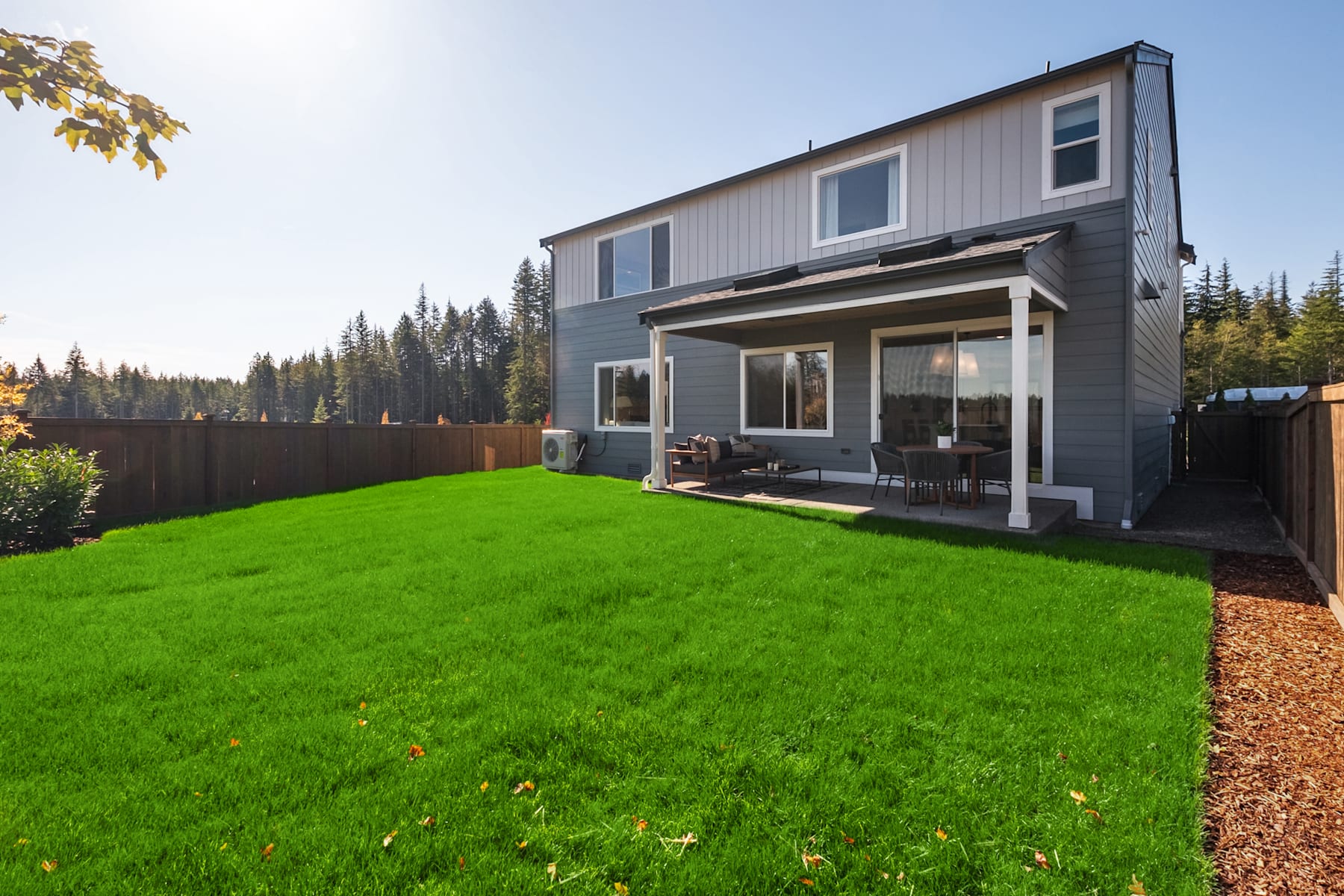 A well-maintained two-story house with a lush green lawn in the foreground, surrounded by a wooden fence and a forested area in the background.