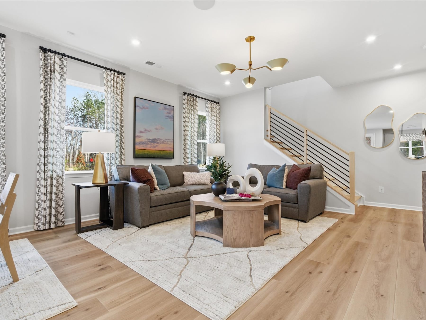 A cozy and modern living room with a plush gray sofa, a wooden coffee table, and a decorative rug on the hardwood floor. The room features large windows, allowing natural light to flood the space, and a stylish chandelier overhead.