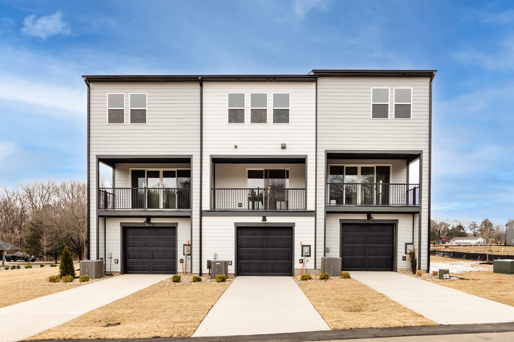 A modern, three-story townhouse with a neutral color scheme, featuring garages and balconies, set against a clear blue sky with some trees in the background.