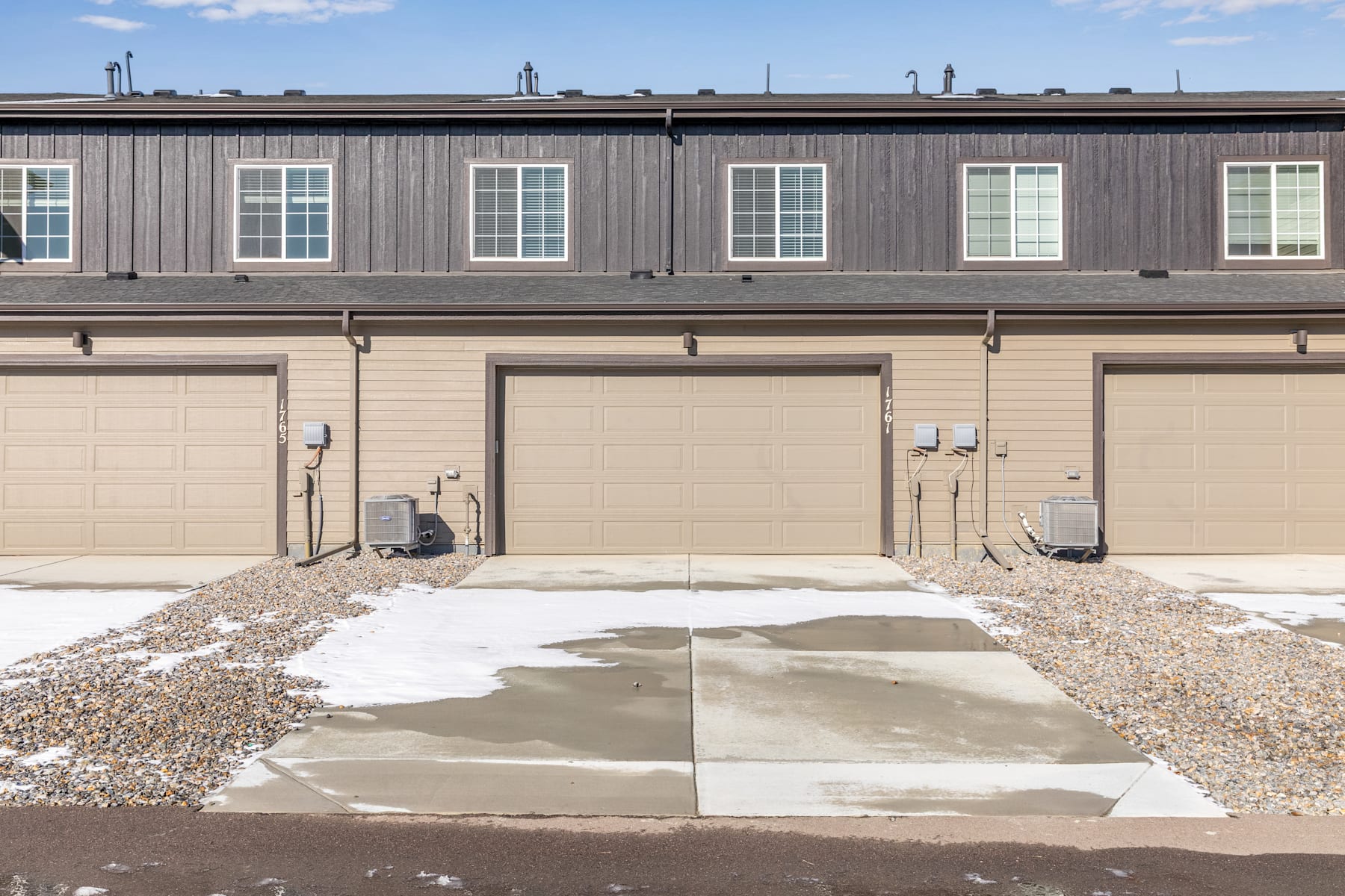 A multi-unit residential building with a gray wooden exterior, featuring several garage doors and windows, stands against a backdrop of a cloudy sky. In the foreground, a concrete walkway leads to the building, surrounded by gravel and snow.