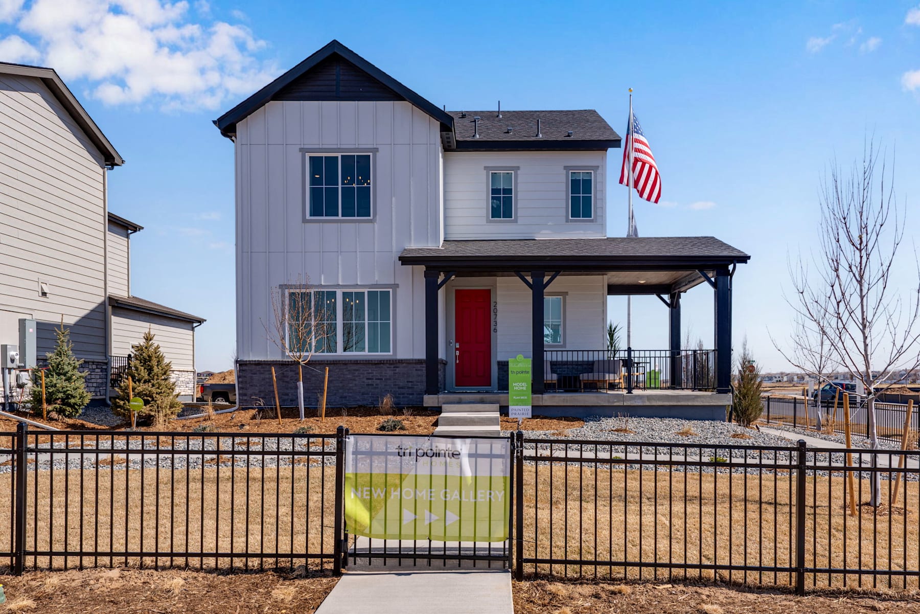A two-story residential house with a porch, American flag, and a fenced yard in the foreground, set against a clear blue sky with some clouds.