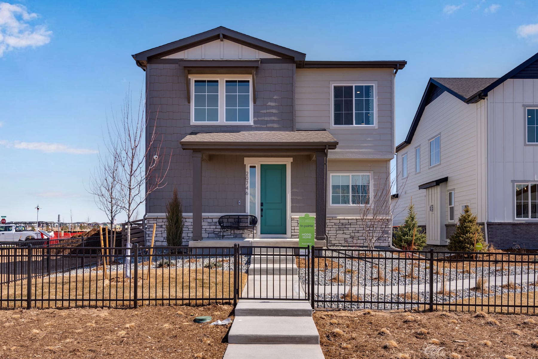 A two-story residential house with a gray exterior, a green front door, and a fenced yard in the foreground, set against a clear blue sky with some clouds.
