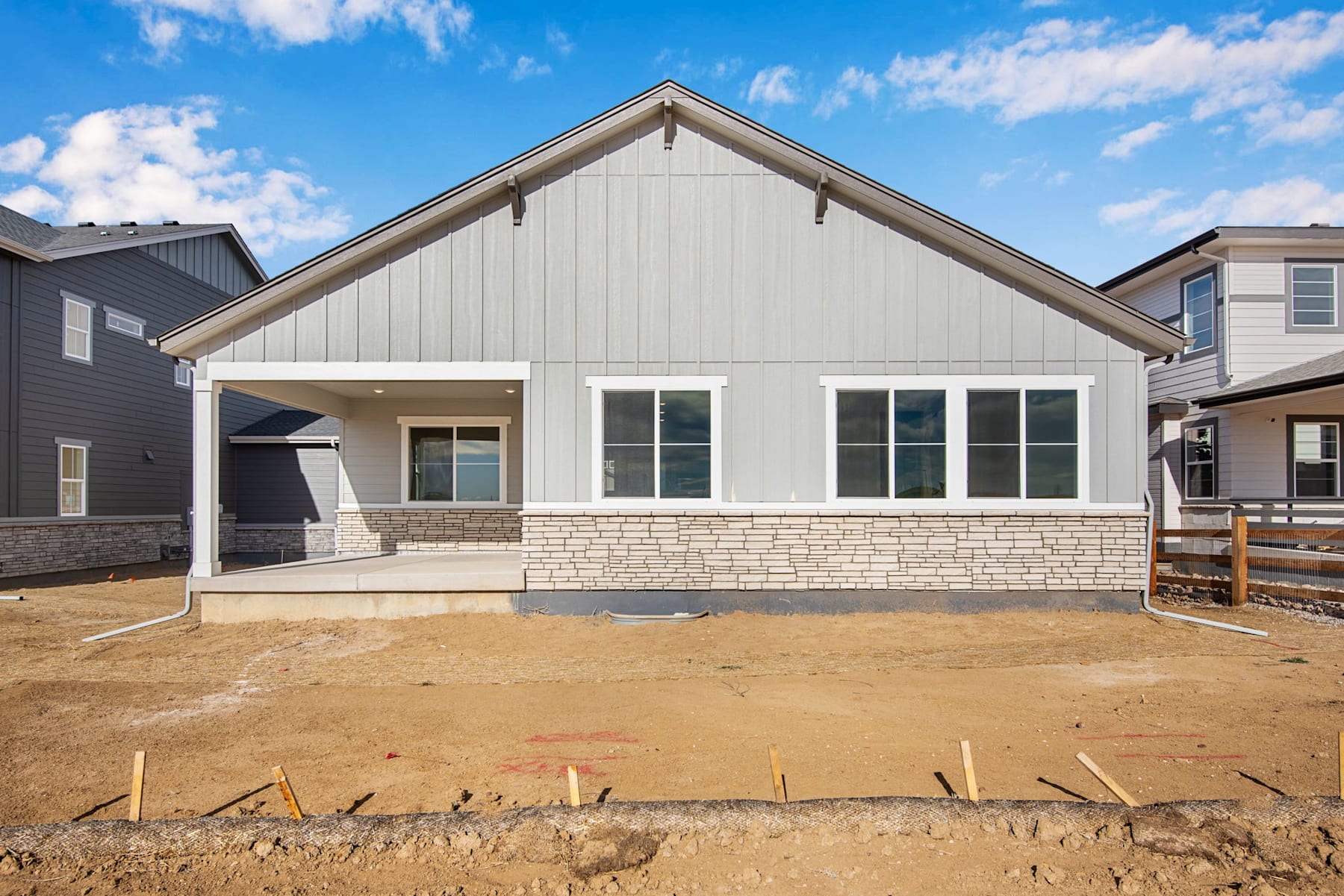 A newly constructed single-story house with a gable roof, white siding, and stone accents, set against a clear blue sky with scattered clouds, surrounded by a dirt construction site in the foreground.