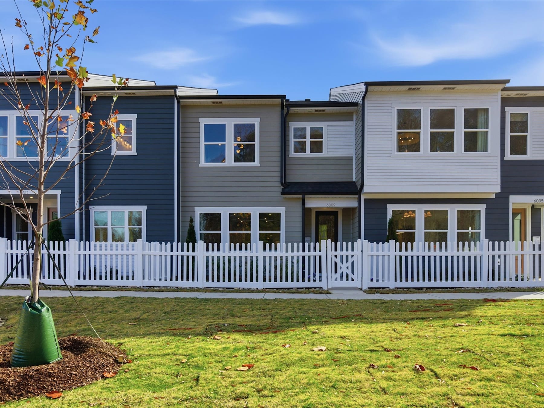 A row of modern, multi-story townhouses with gray and white siding, surrounded by a grassy lawn and a white picket fence in the foreground.