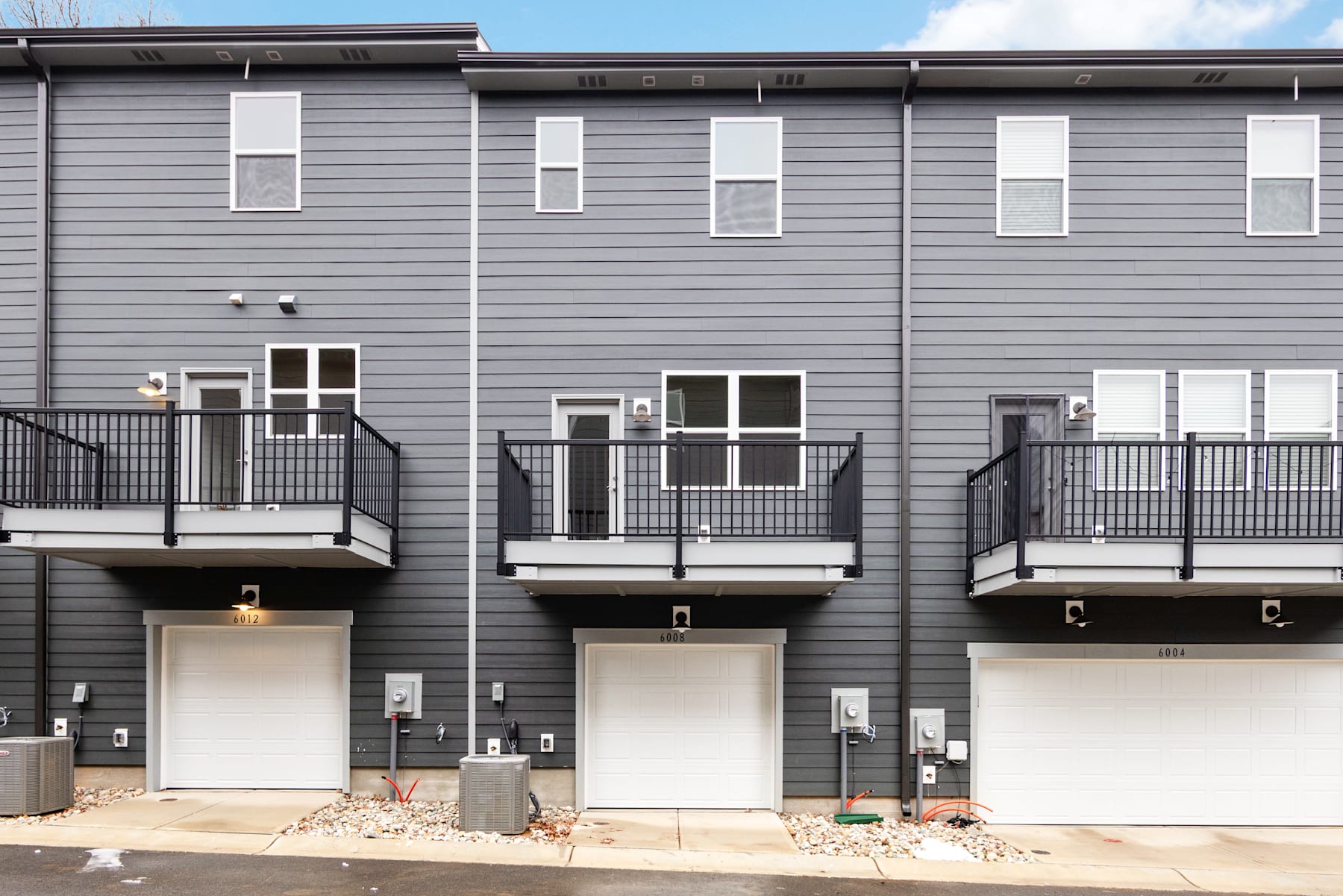 A multi-story residential building with gray siding, balconies, and garage doors in the foreground, set against a clear blue sky in the background.