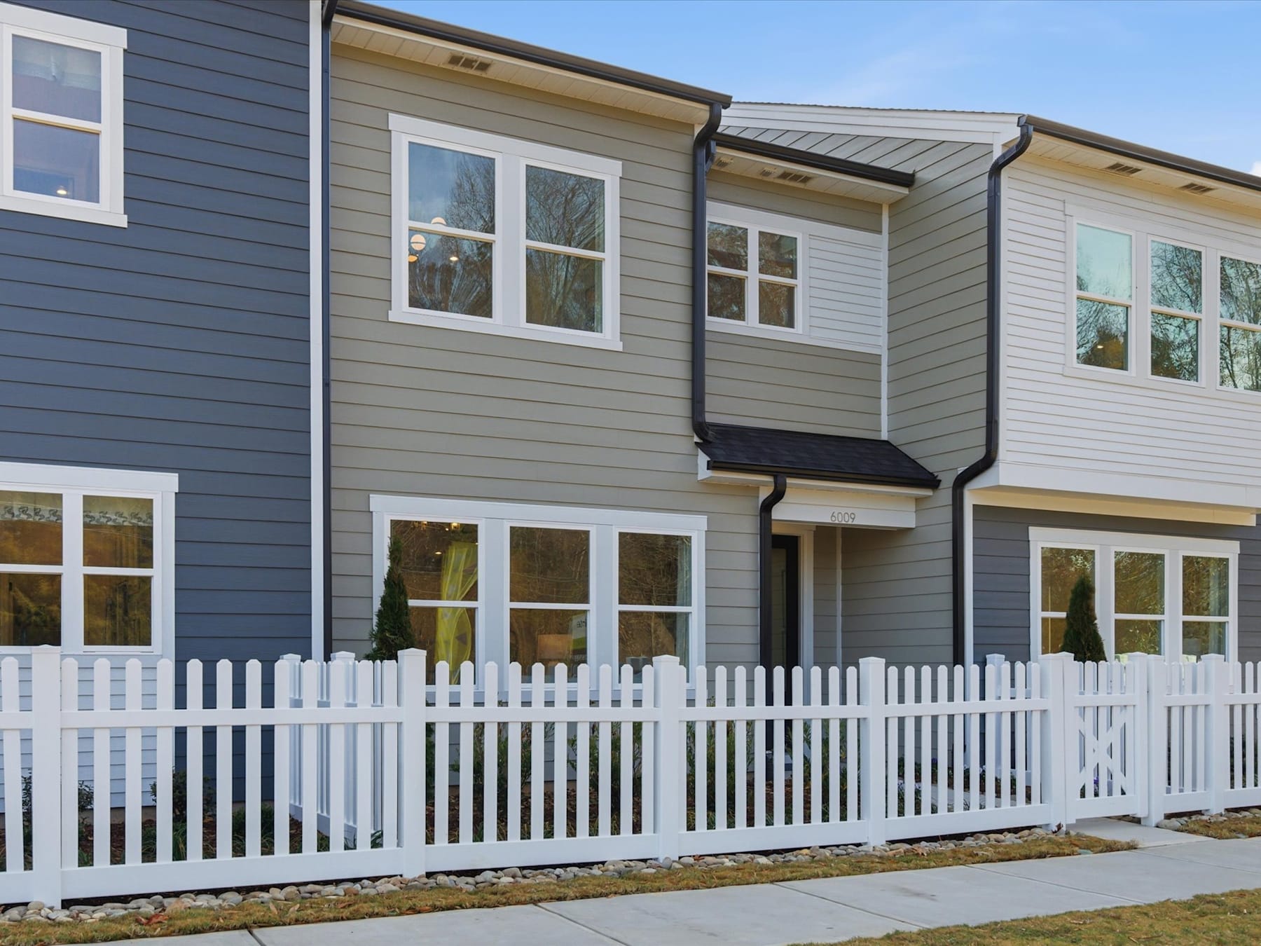 A two-story residential building with gray siding and a white picket fence in the foreground, set against a clear blue sky.