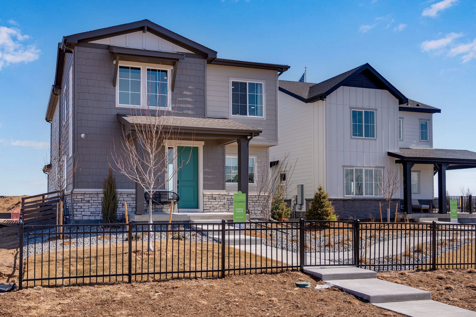 A two-story residential house with a gray exterior, a green front door, and a wrought-iron fence surrounding the front yard, set against a clear blue sky with some clouds.