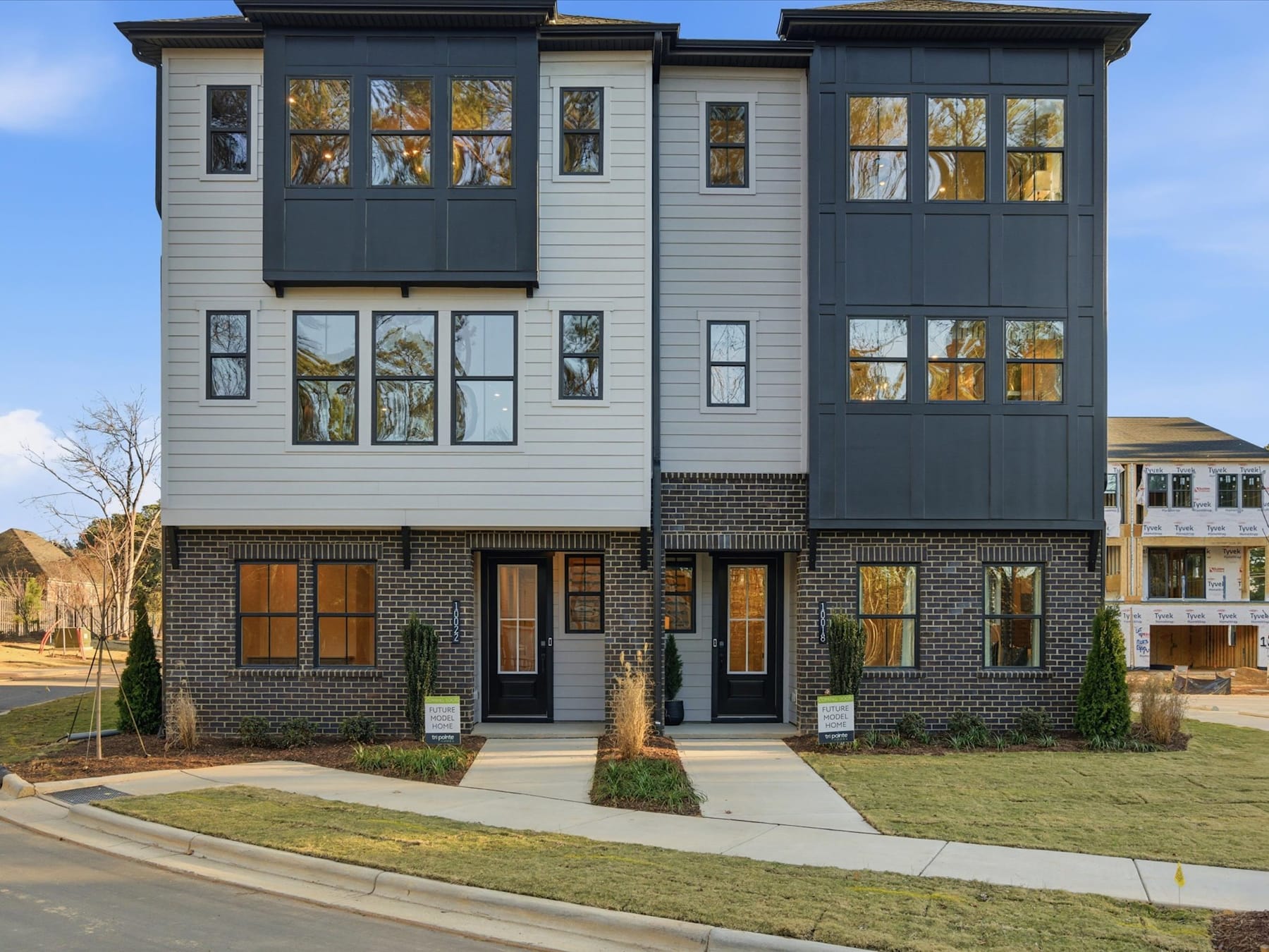 A multi-story residential building with a mix of gray siding and brick exterior, surrounded by a grassy lawn and other buildings in the background.