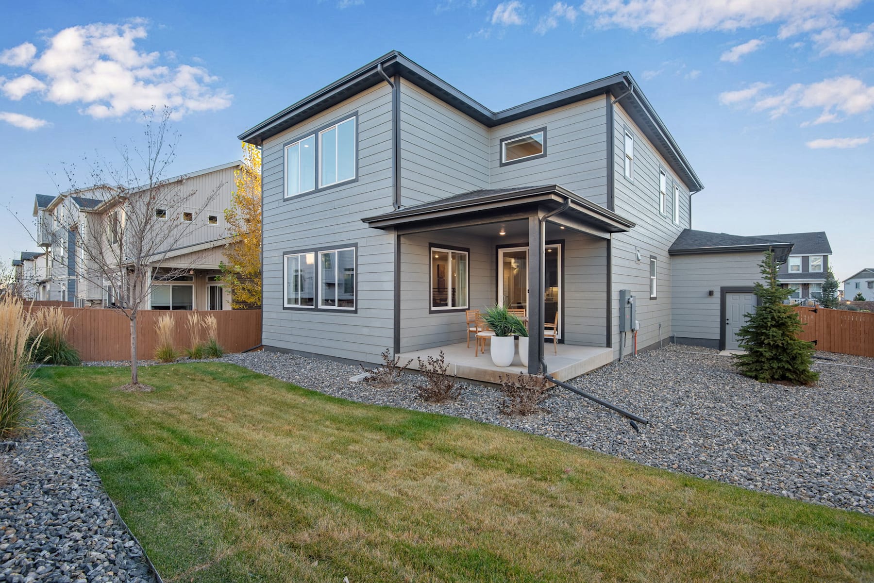 A two-story gray house with a gravel driveway and a well-maintained lawn in the foreground, set against a backdrop of a clear blue sky with scattered clouds.