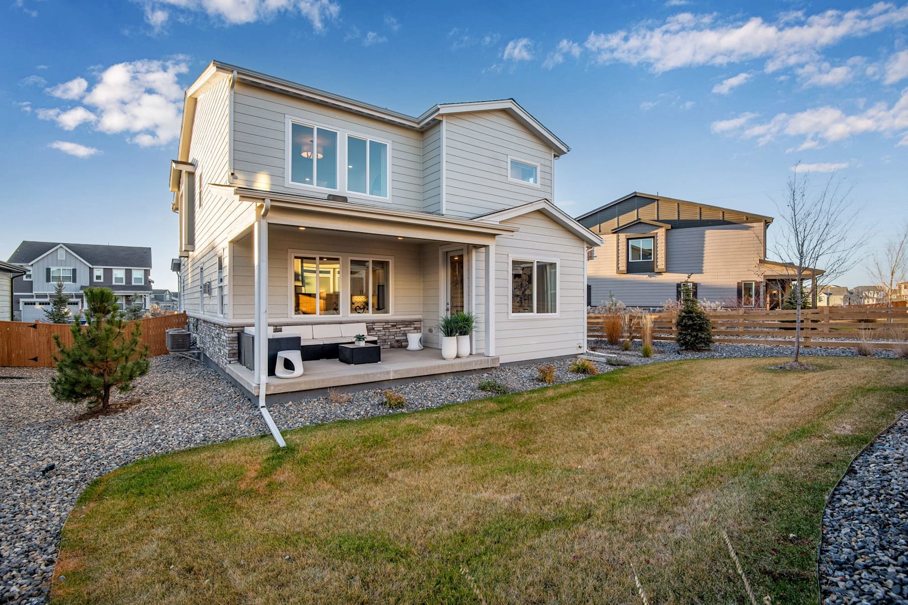 A two-story residential house with a porch, surrounded by a grassy yard and other houses in the background, set against a blue sky with fluffy white clouds.