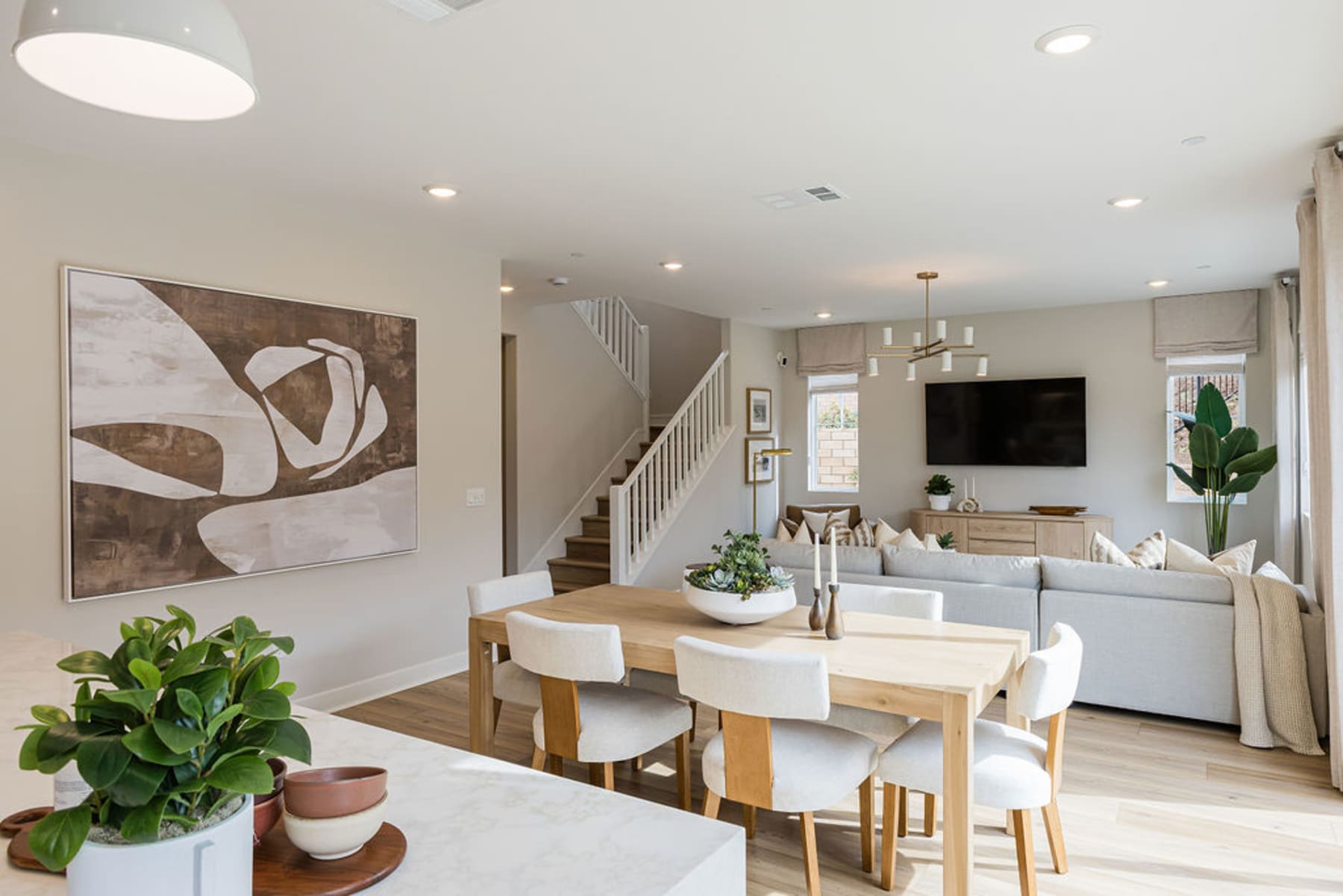 A modern and minimalist living room with a dining area, featuring a large abstract artwork, potted plants, and a sleek, neutral-toned interior design.