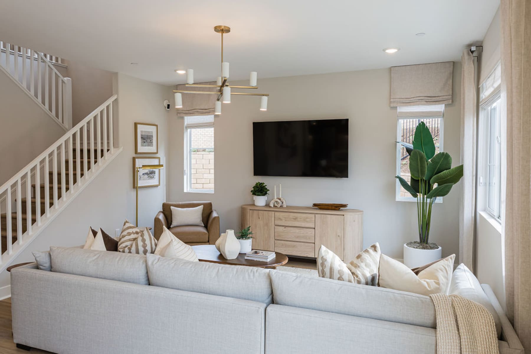 A cozy and inviting living room with a large gray sofa, a wooden dresser, and a large TV mounted on the wall, complemented by decorative plants and artwork.