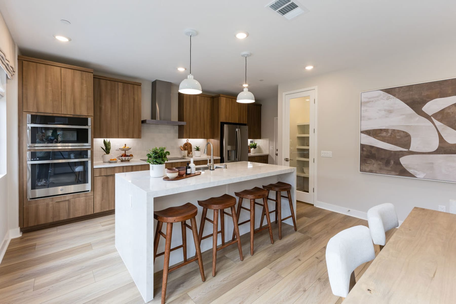 A modern and spacious kitchen with a large island, wooden cabinets, and pendant lights, set against a neutral-toned living area with a large abstract artwork on the wall.