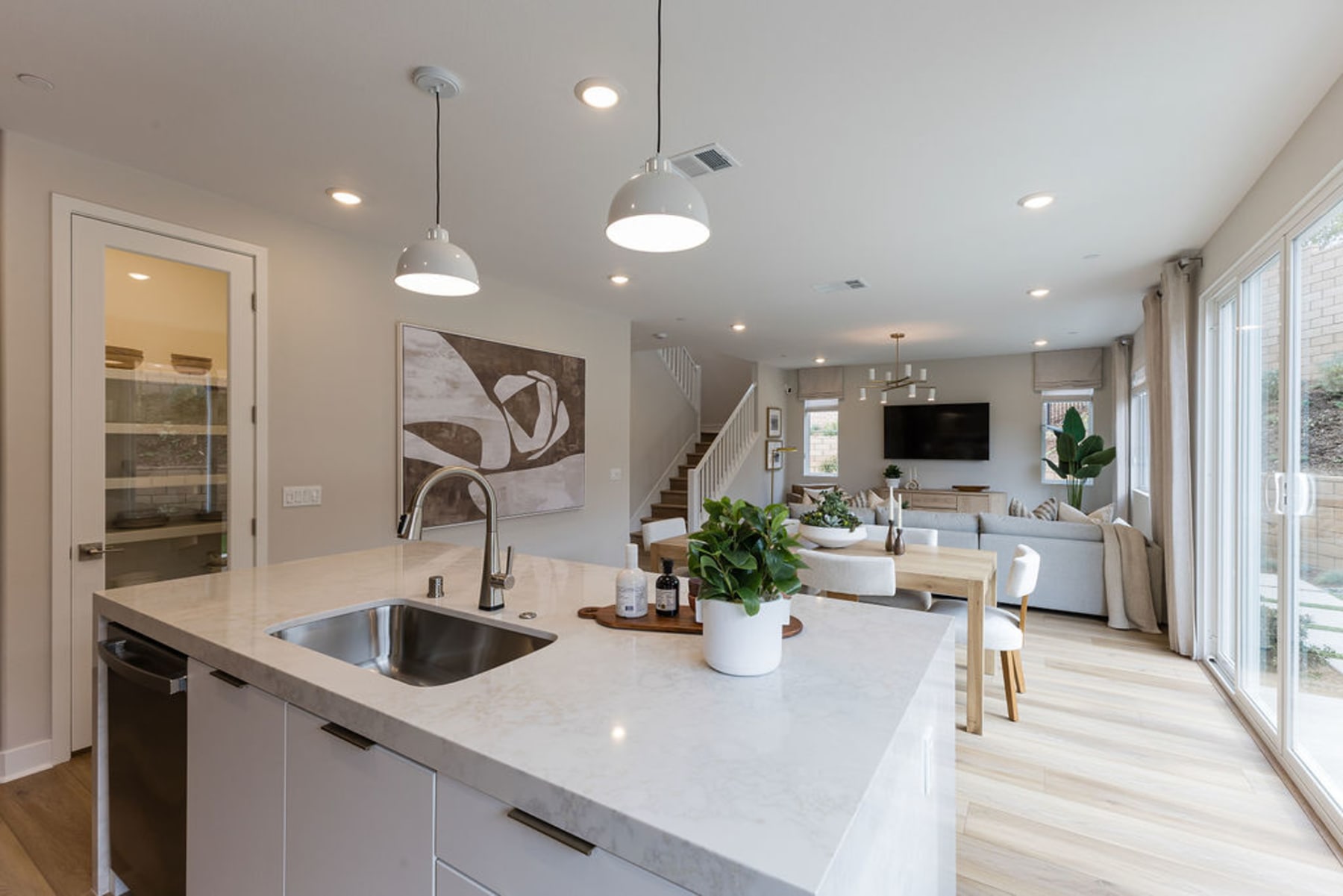 A modern, open-concept kitchen and living room with white countertops, pendant lighting, and a large window providing natural light.