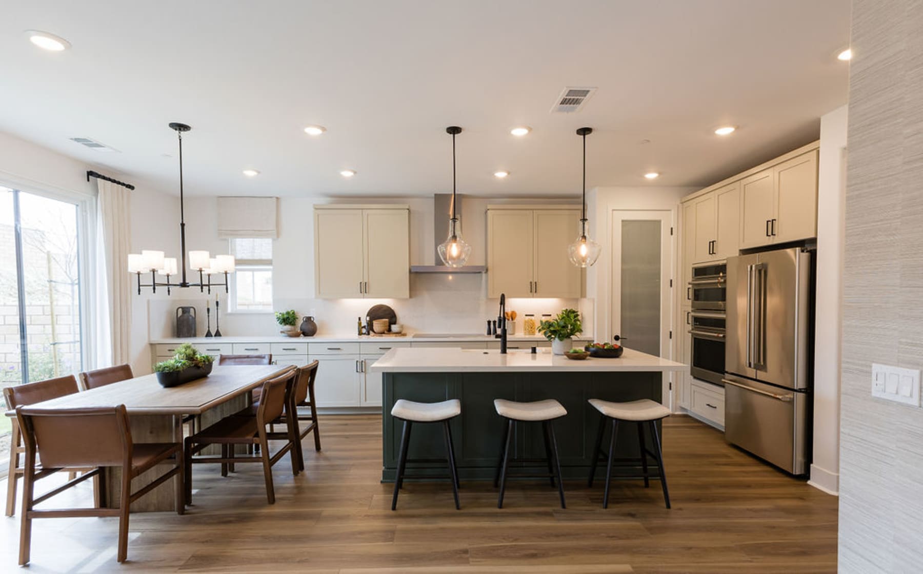 A modern and spacious kitchen with light-colored cabinets, a large island with bar stools, and a dining area with a wooden table and chairs, all set against a backdrop of hardwood floors and large windows.