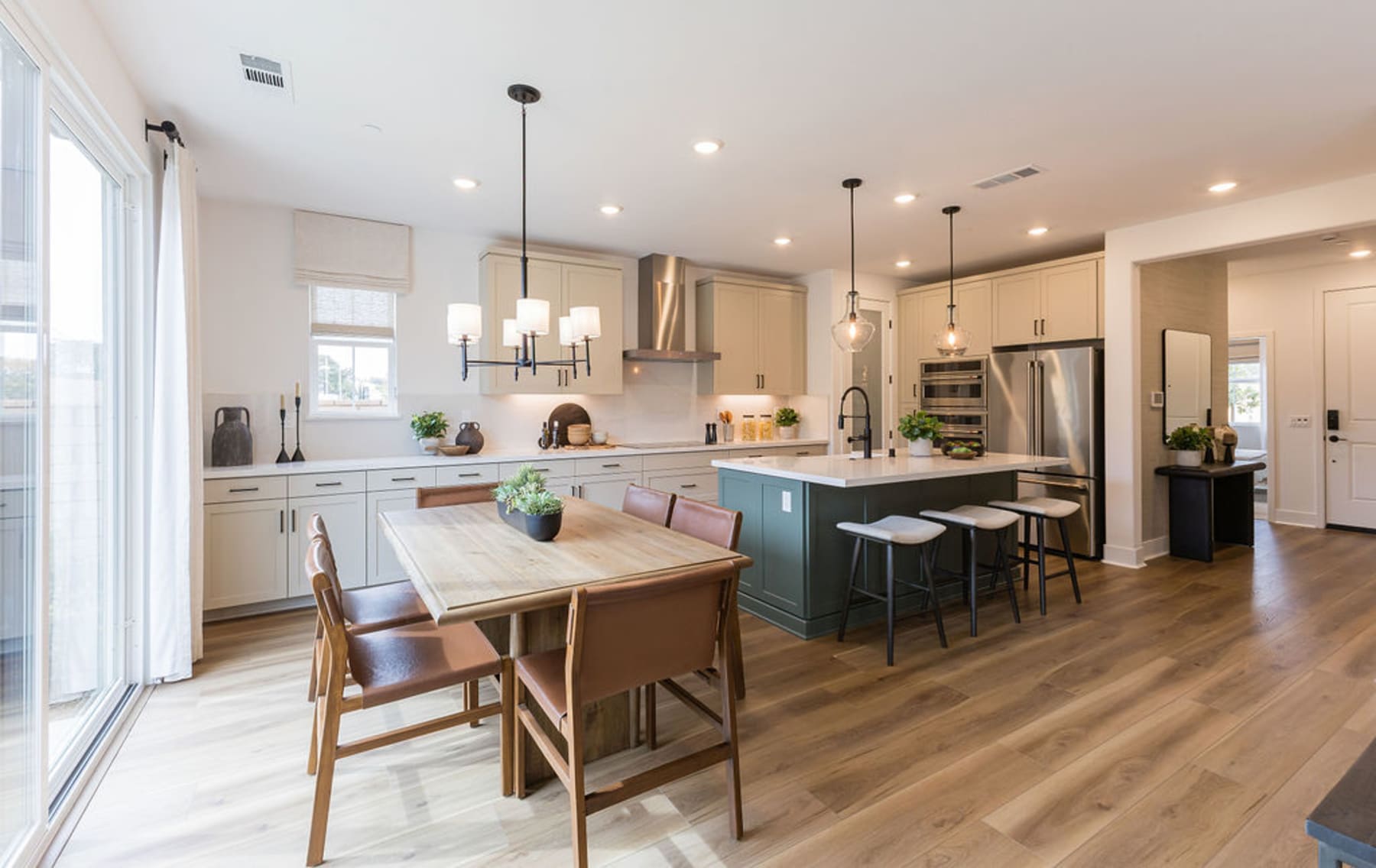 A modern and spacious kitchen with a wooden dining table, chairs, and a kitchen island with stools, surrounded by white cabinets, hardwood floors, and pendant lighting.