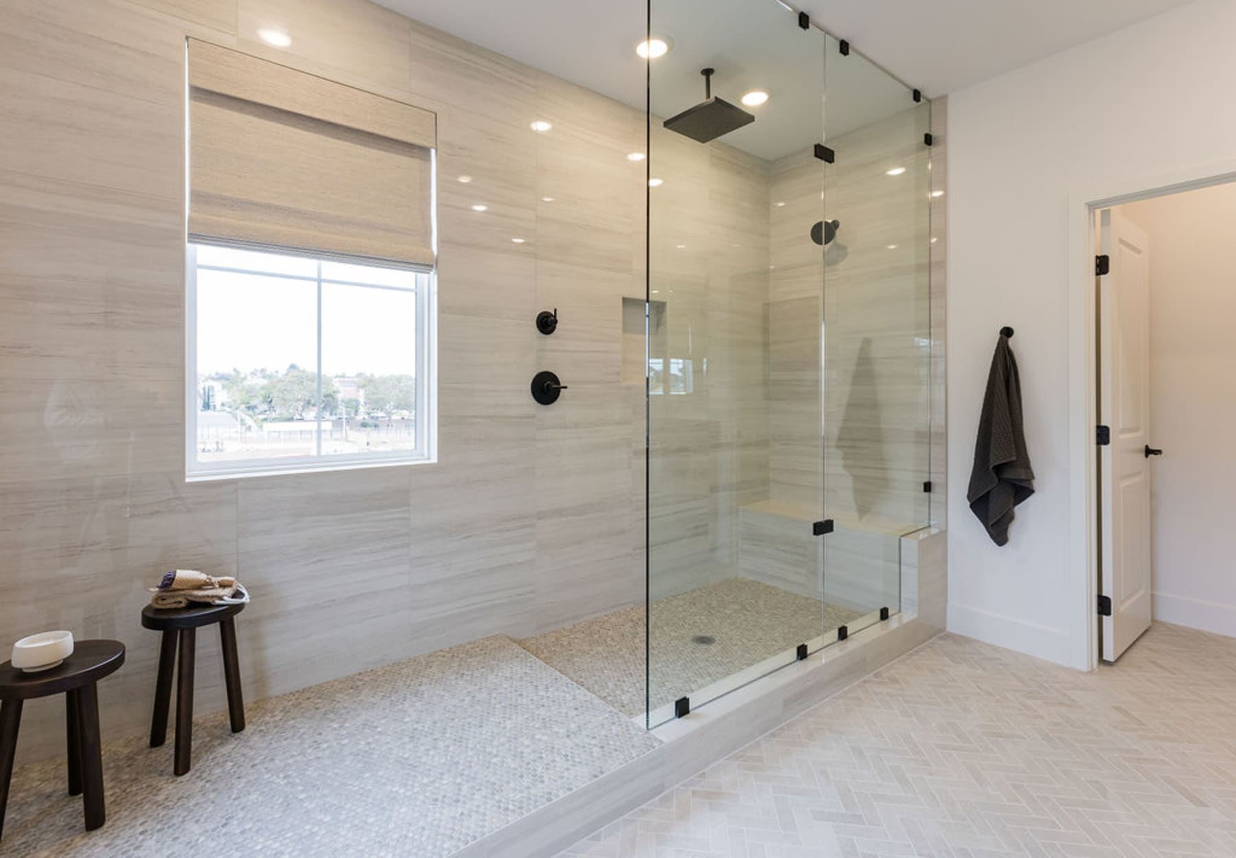 A modern, well-lit bathroom with a large glass shower enclosure, a window providing natural light, and wooden stools for seating.