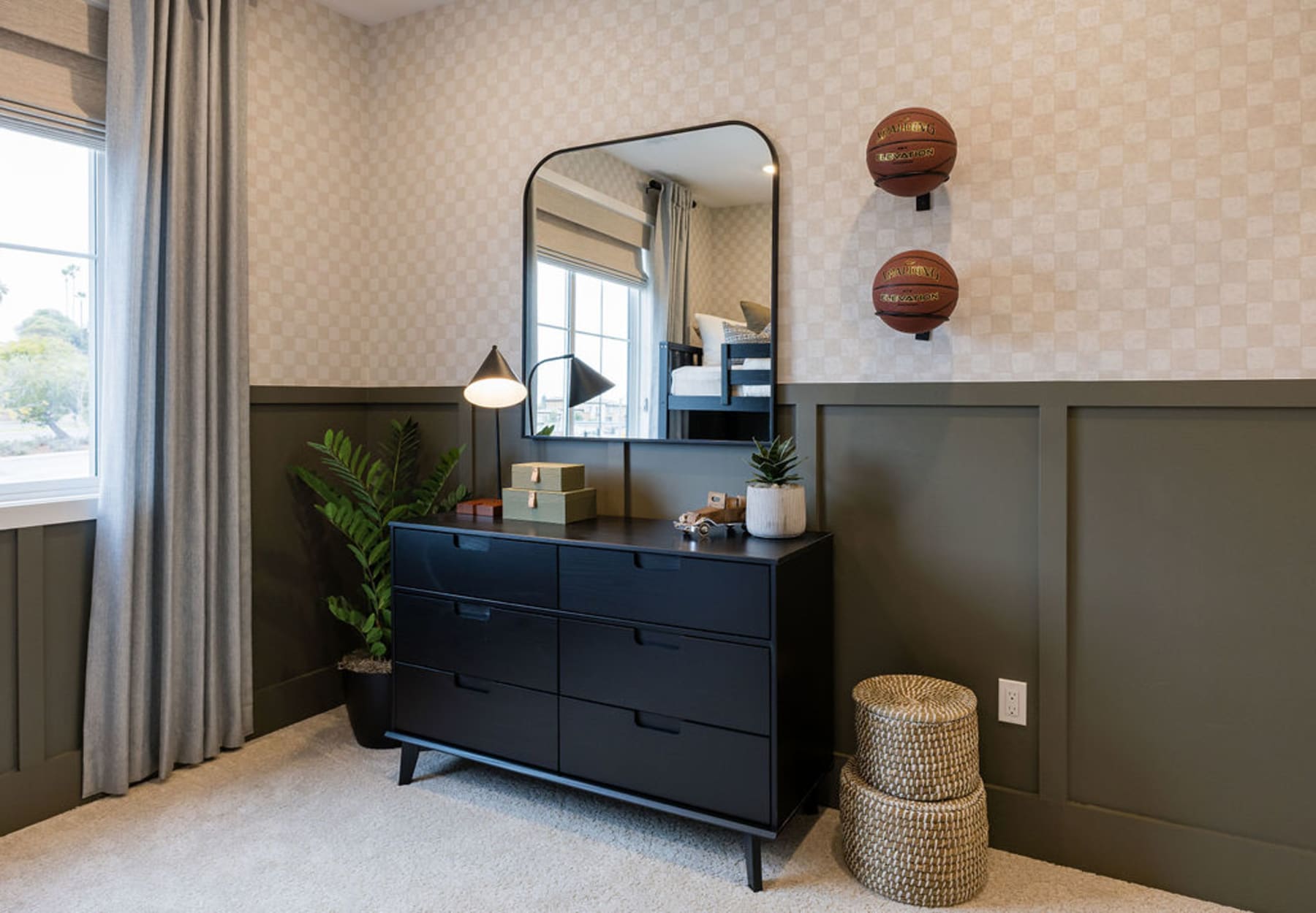 A modern and stylish bedroom interior with a black dresser, a large mirror, and decorative elements such as plants and woven baskets.