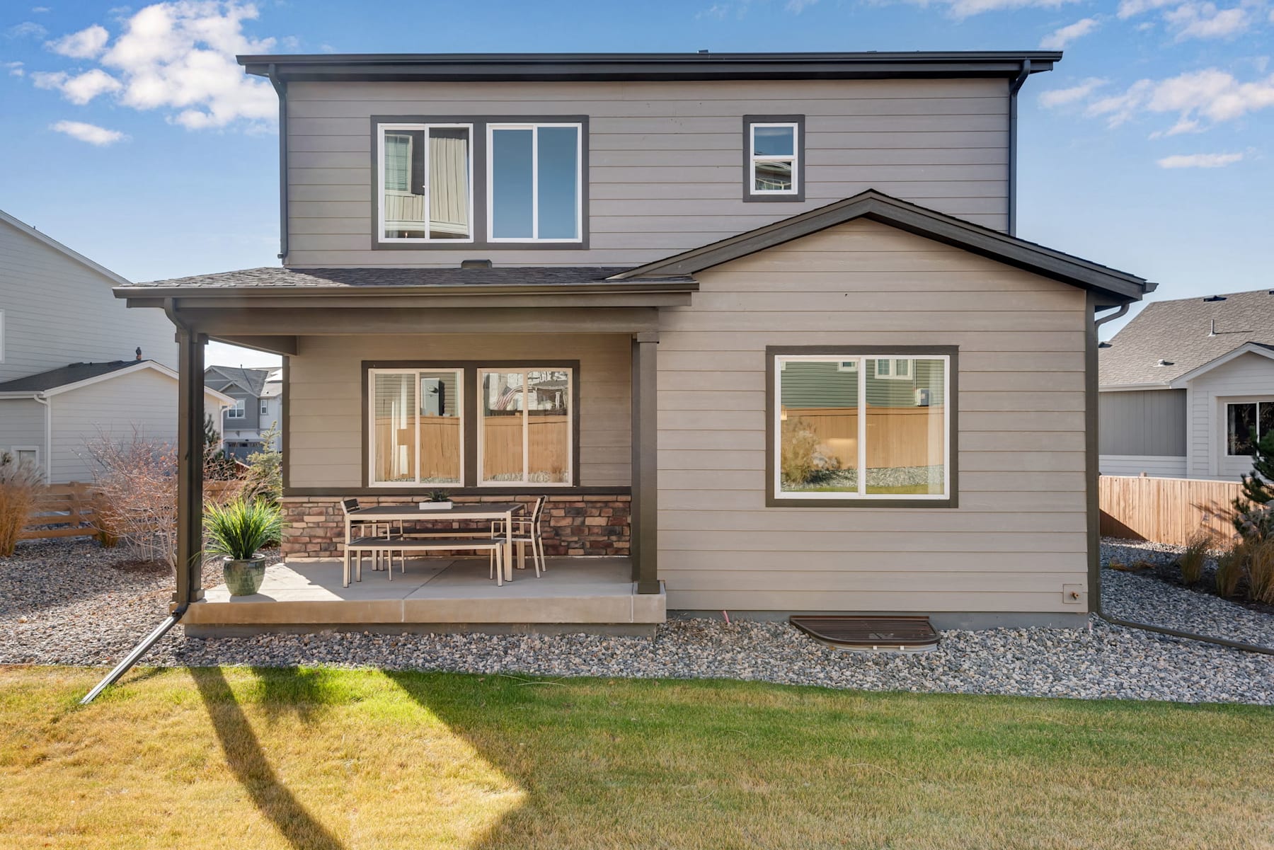 A two-story residential house with a gray exterior, a porch, and a gravel driveway surrounded by a grassy lawn and a clear blue sky with scattered clouds.