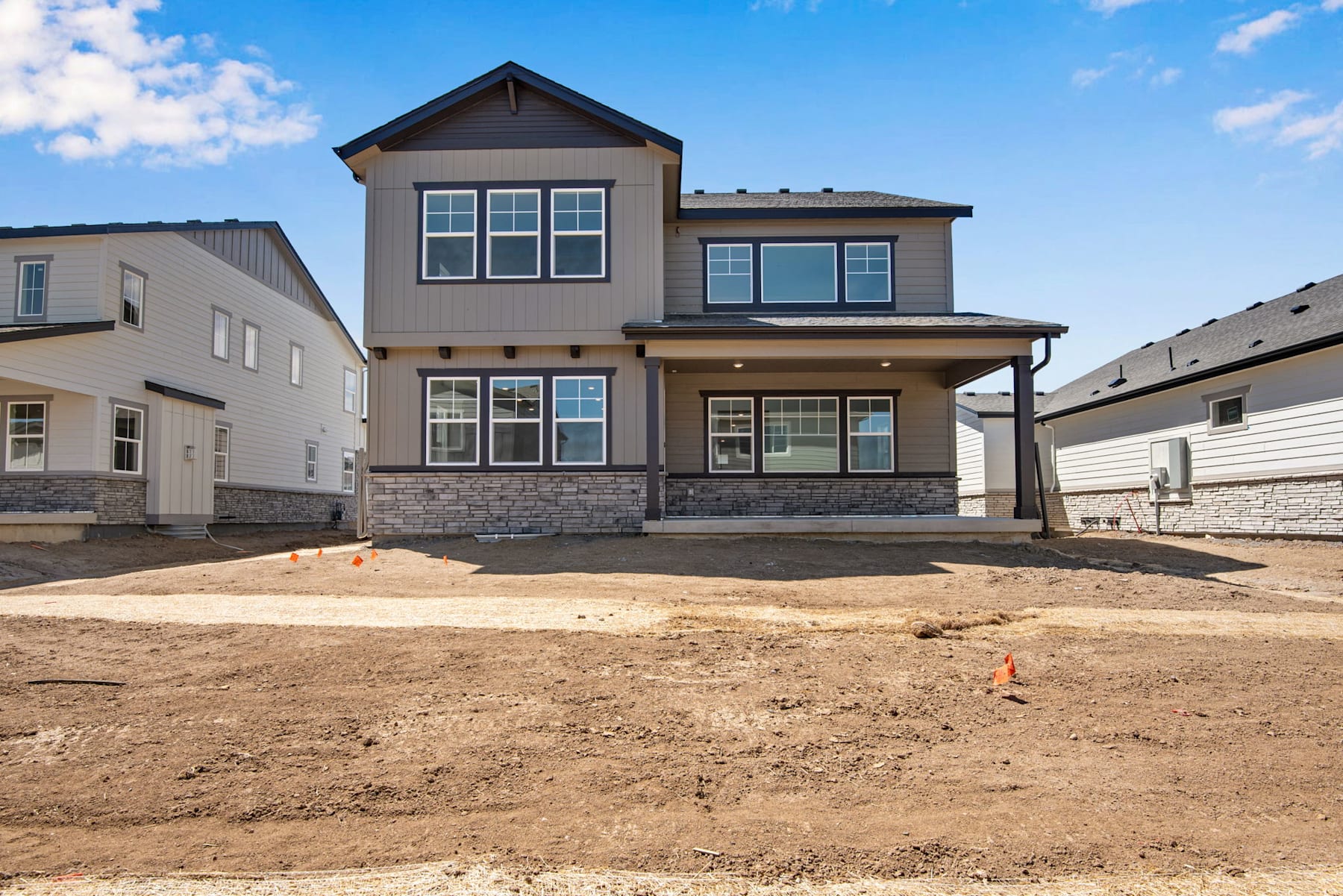 A newly constructed two-story residential house with a stone exterior, surrounded by a dirt construction site and other houses in the background.