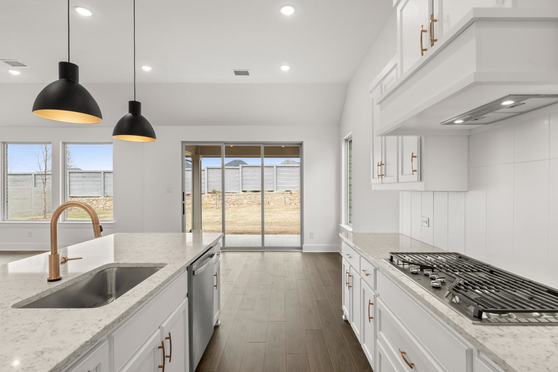 A modern, well-lit kitchen with white cabinets, a stainless steel sink, and pendant lights hanging above a countertop. The kitchen opens up to a bright, airy space with large windows providing a view of the outdoors.