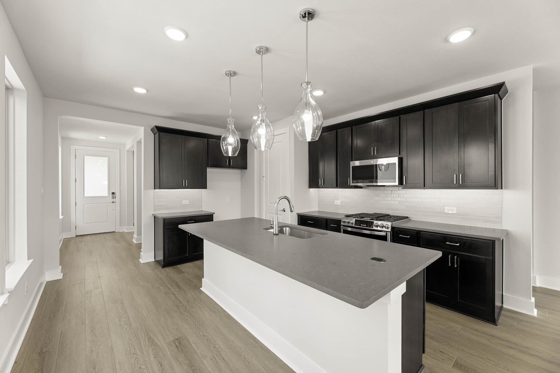 A modern, open-concept kitchen with dark cabinets, a gray countertop, and pendant lighting fixtures, set against a light-colored hardwood floor.