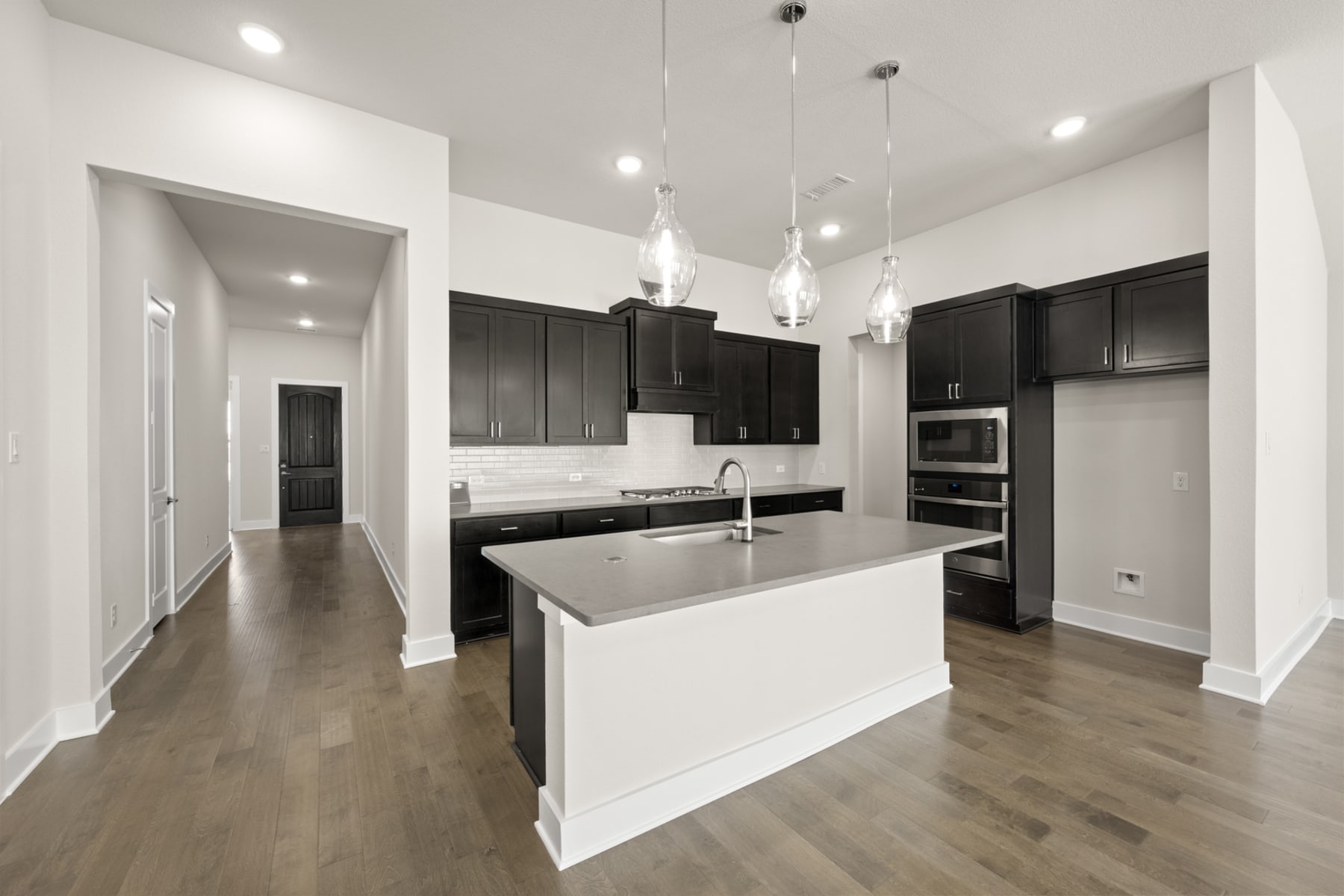 A modern, open-concept kitchen with dark cabinets, a white island, and pendant lighting fixtures, set against a backdrop of hardwood floors and a hallway leading to another room.