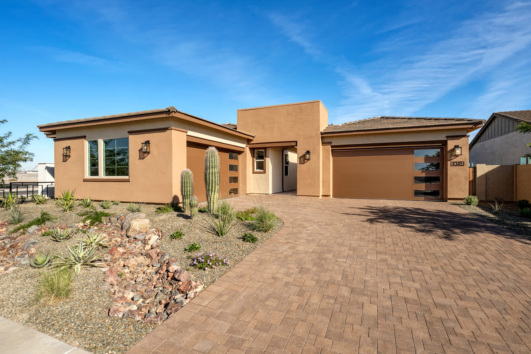 A single-story, stucco-clad house with a tiled roof sits on a paved driveway surrounded by desert landscaping, against a backdrop of a clear blue sky with wispy clouds.