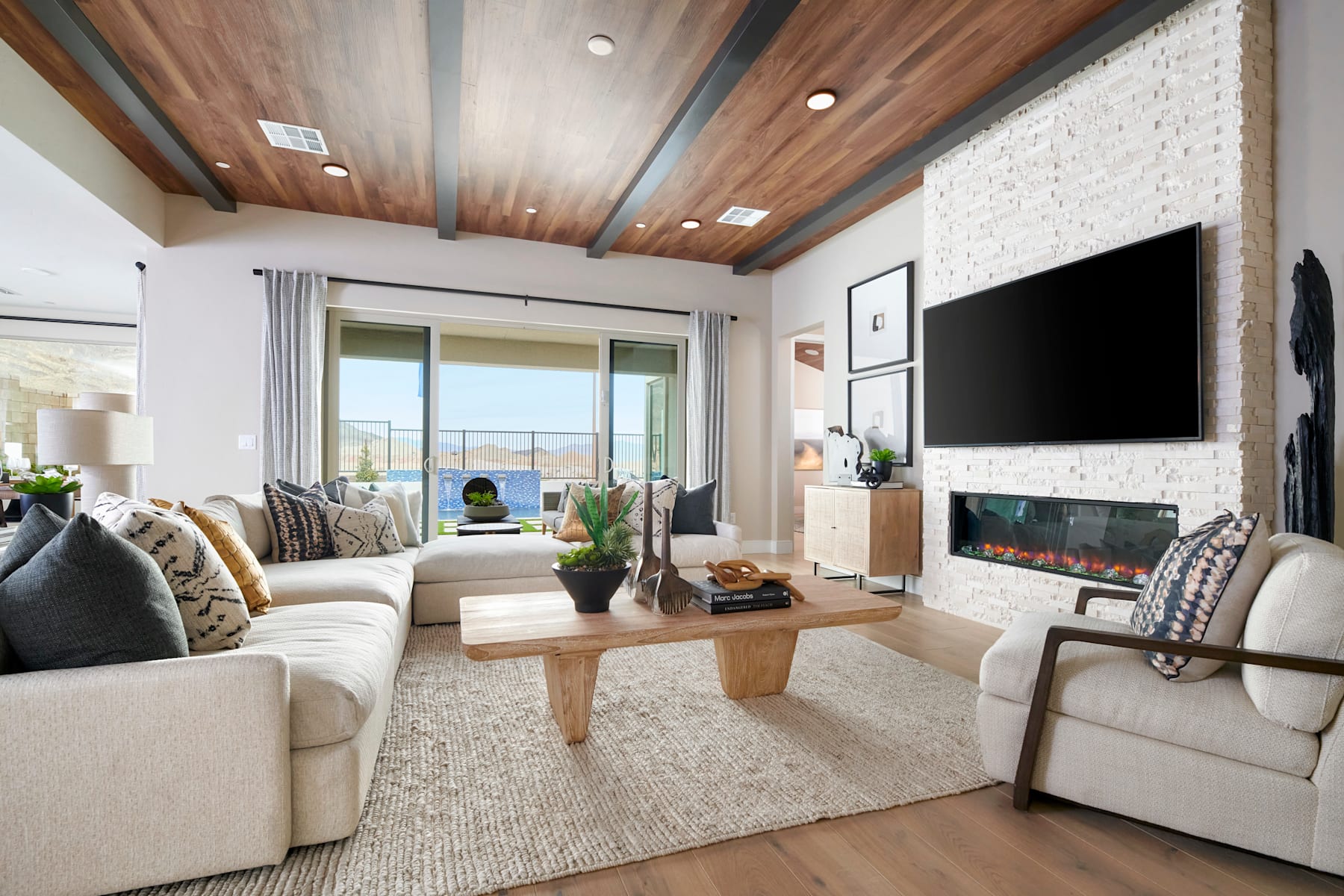 A cozy and inviting living room with wooden ceiling beams, a fireplace, and large windows overlooking a scenic outdoor view.