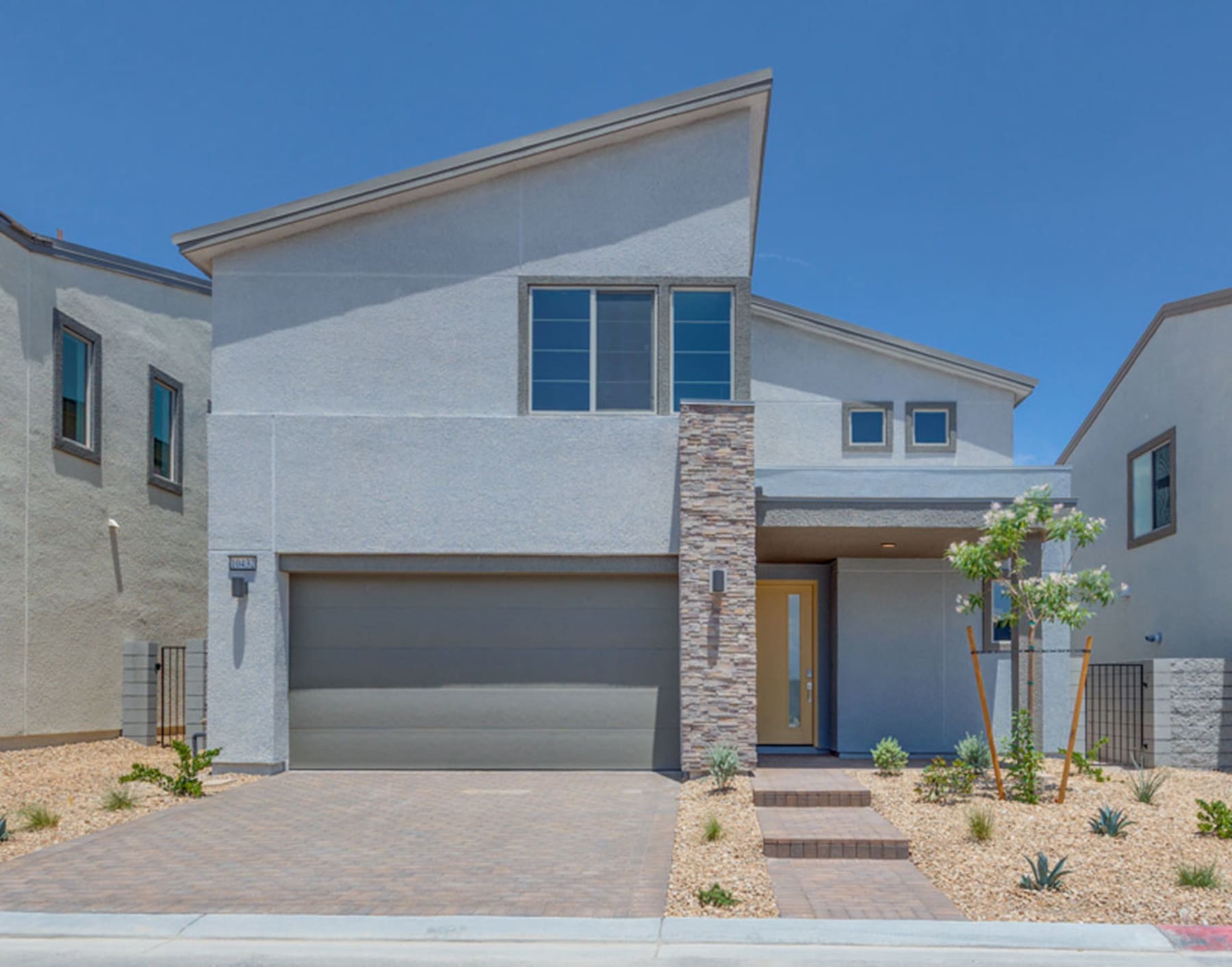 A modern, two-story house with a gray exterior, stone accents, and a garage door stands in a well-landscaped yard with a paved driveway and potted plants.