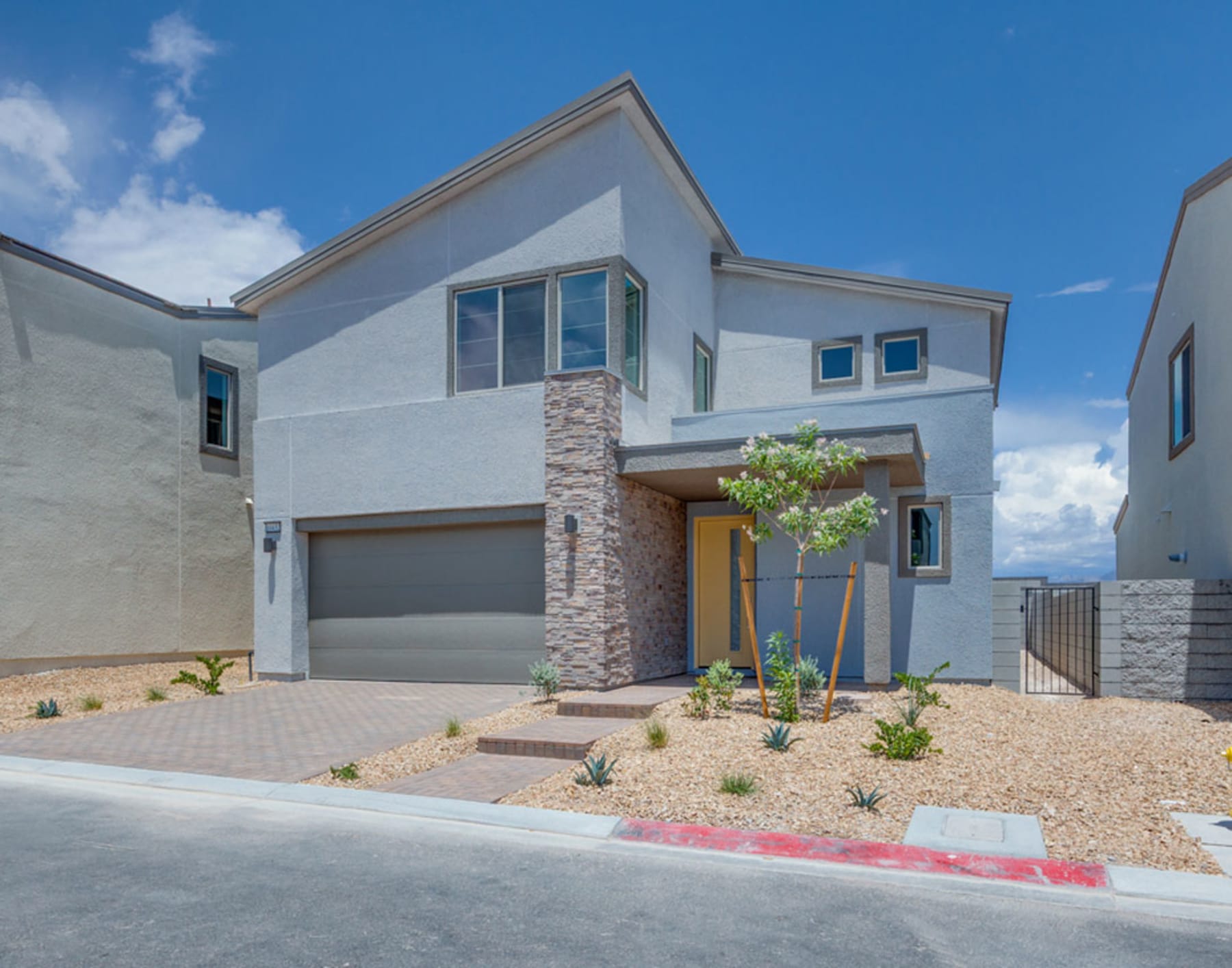 A modern two-story house with a garage, surrounded by a landscaped yard with desert plants and a paved driveway, set against a clear blue sky.