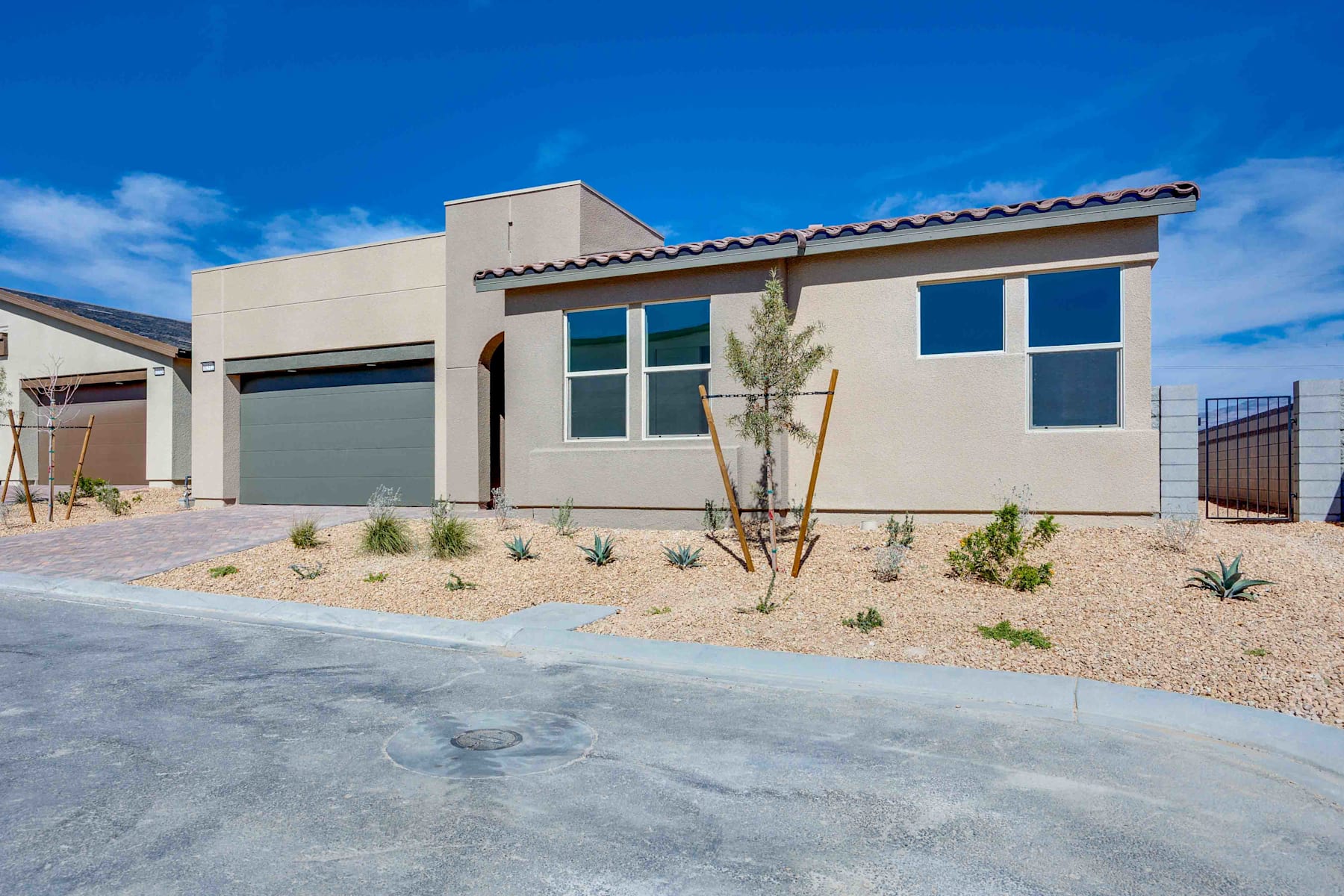 A modern, single-story beige building with a tiled roof stands in the foreground, surrounded by a gravel driveway and desert landscaping against a clear blue sky.