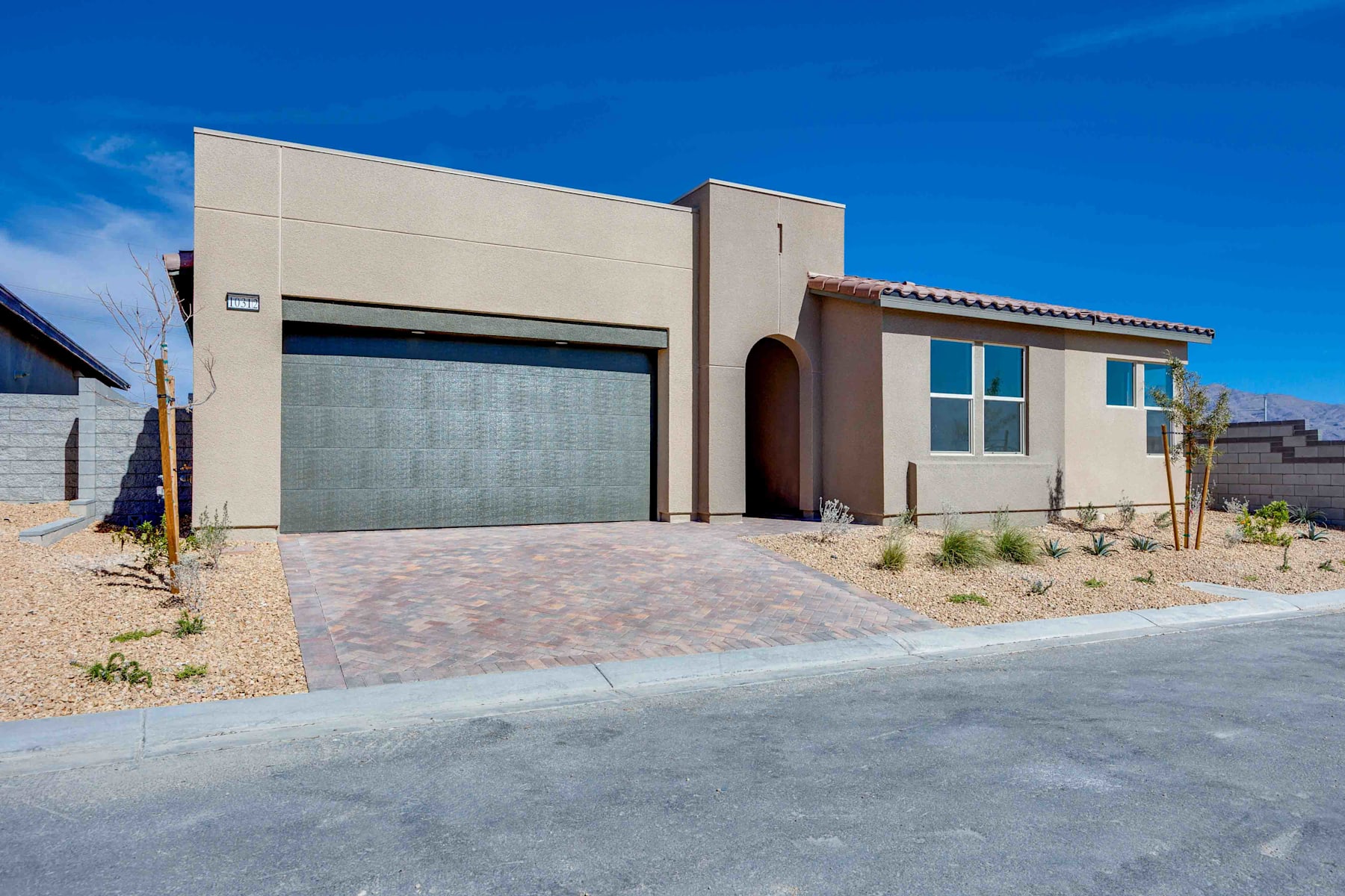 A modern, single-story house with a garage door and a paved driveway, set against a clear blue sky with some clouds.