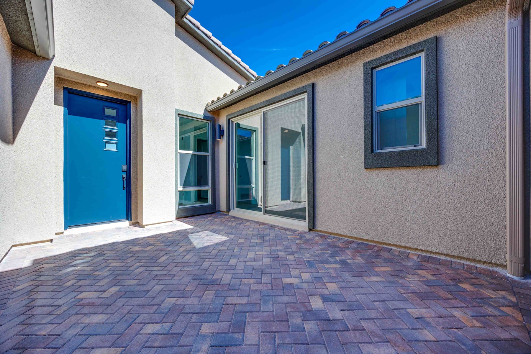 A brick-paved walkway leads to a modern, multi-windowed building with a vibrant blue door, set against a clear blue sky.